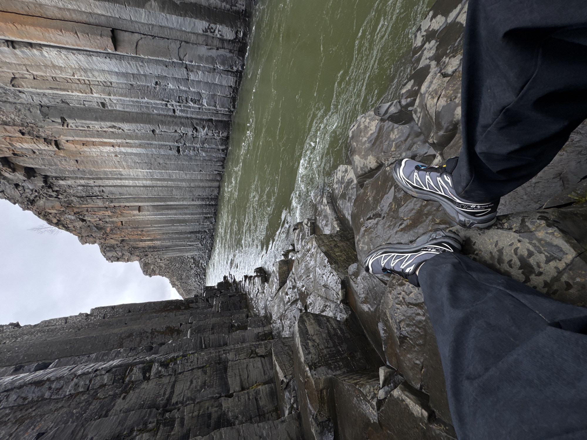 The image presents a first-person perspective looking down into a dramatic natural gorge, showcasing a person's feet positioned precariously on a rocky ledge. The scene is dominated by striking columnar basalt formations that line the sides of a deep chasm. On the left side of the frame, towering grey and dark basalt columns rise vertically, displaying distinct horizontal striations and varying shades of grey, with some brownish tones visible on weathered surfaces.
A murky green river flows rapidly through the bottom of the gorge, its surface agitated with ripples and white streaks of foam, indicating strong currents. On the right side, the person's lower legs and feet are visible. They are wearing dark pants, possibly waterproof, and dark grey hiking shoes with white accents and yellow details. The brand "Salomon" is clearly visible on the side of the shoes, along with "XT-6" and "GORE-TEX" branding, indicating specialized outdoor footwear. The rocks immediately around the person's feet appear wet and slick, suggesting recent rain or spray from the river.
Above the gorge, a pale, overcast sky is visible, contributing to the diffused, cool lighting of the scene. The overall impression is one of rugged natural beauty and adventure, with the basalt columns being a strong geological indicator of a volcanic region, consistent with the location hint of Iceland. The perspective emphasizes the depth and scale of the gorge.