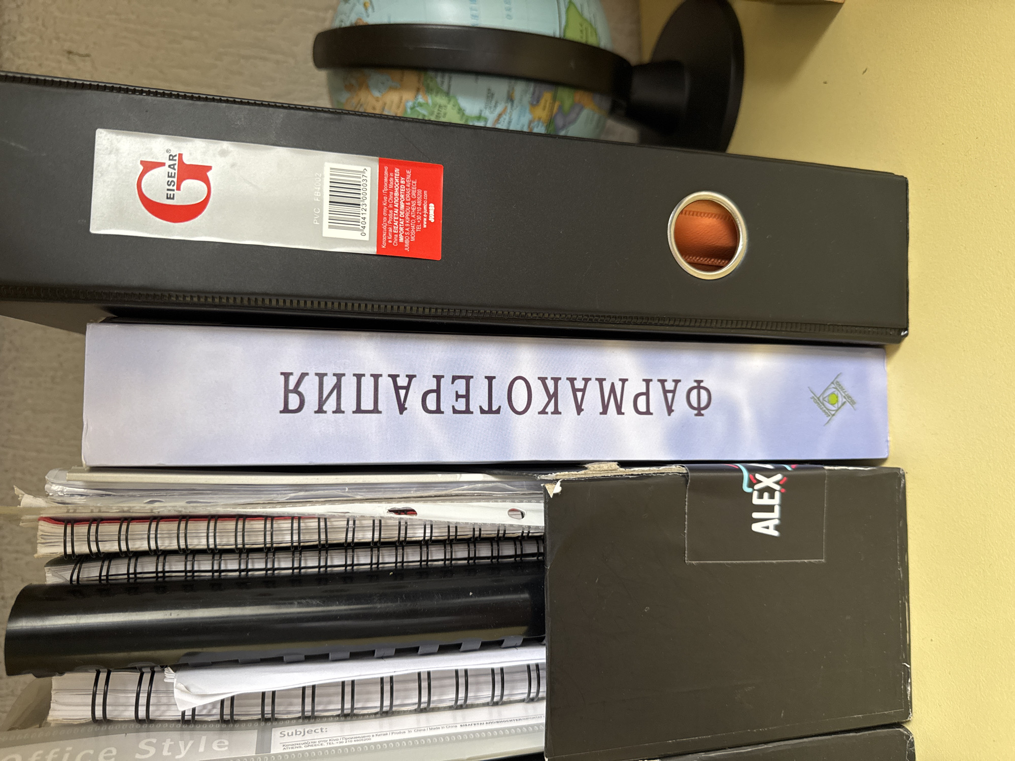 The image displays a shelf with several binders and notebooks. On the far left, partially visible, is a black binder with white text that includes the word "ALEX". To its right is a light blue binder with dark purple text that reads "ФАРМАКОТЕРАПИЯ" in Cyrillic. Further to the right is a large black binder with a silver ring hole and a red and white label featuring the "EISEAR" logo. The background shows a portion of a globe. The scene is on a yellow surface, likely a desk or shelf. There are no people or animals present. The overall impression is that of a home or office setting, with the binders and notebooks organized on a shelf. The city of Smolyan, Bulgaria, is not depicted or referenced in the image.