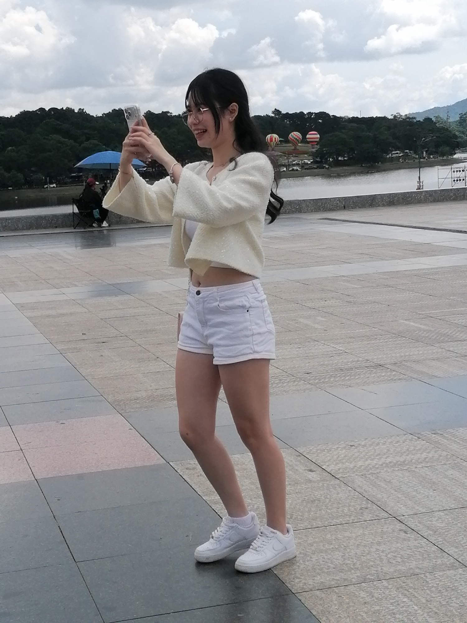 A young woman with dark hair and glasses is standing outdoors on a paved area, holding a cell phone to take a selfie. She is wearing a cream-colored, sequined jacket over a white crop top and white denim shorts. She is also wearing white sneakers. In the background, there is a body of water, a line of trees, and mountains. Several hot air balloons are visible in the distance. The sky is cloudy. To the left of the woman, a person sits on a chair under a blue umbrella. The scene appears to be at Xuan Huong Lake in Da Lat, Vietnam.