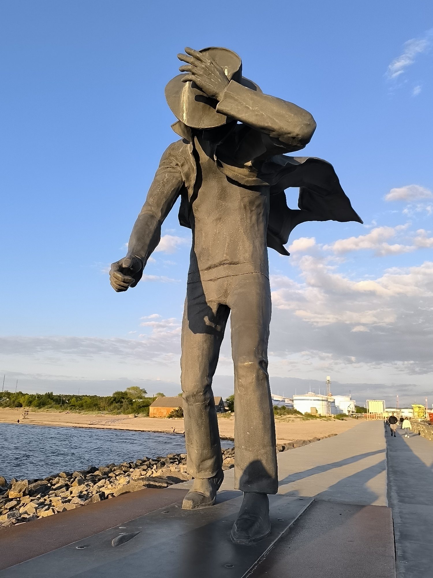 A low-angle shot captures a dark-colored statue of a man standing prominently by a waterfront. The statue depicts a male figure in a jacket and trousers, with his right hand holding a hat to shield his face or eyes, and his left arm bent with a clenched fist. His jacket and scarf appear to be blown by the wind, suggesting dynamic movement or a strong breeze. The statue stands on a dark, textured base next to a paved walkway.
To the left of the statue, a body of water with gentle waves meets a rocky shoreline. Beyond the rocks, a sandy beach stretches into the distance, backed by green vegetation and a small, reddish-brown building.
To the right, the paved walkway extends far into the background, where two individuals are visible walking away from the viewer. Long shadows, cast by the statue and the distant walkers, stretch across the walkway, indicating a low sun angle, likely late afternoon or early morning. In the far background, several white industrial-style tanks and a communication tower can be seen.
The sky is a vibrant blue with scattered white, fluffy clouds. The overall lighting is warm, enhancing the sense of the golden hour. No visible text is present in the image. The scene suggests a coastal or lakeside setting, with the statue appearing to gaze out at the water, perhaps contemplating or braving the elements.