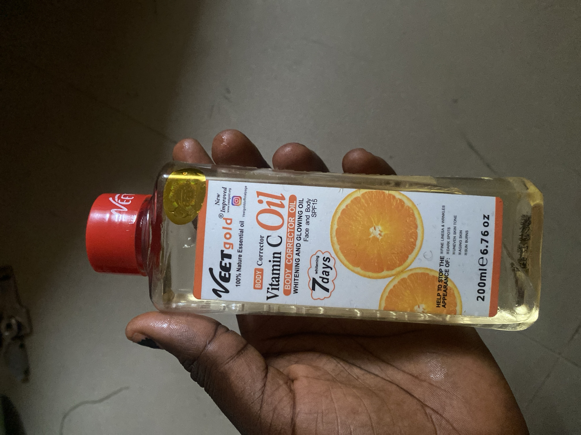 The image shows a close-up of a person's hand holding a bottle of "Veet Gold Vitamin C Oil." The bottle is rectangular with a red cap and contains a clear, yellowish oil. The label on the bottle prominently features the brand name "Veet Gold" and states "BODY CORRECTOR OIL WHITENING AND GLOWING OIL FACE AND BODY SPF15." It also lists benefits such as helping to stop the appearance of fine lines, wrinkles, dark spots, uneven skin tone, aging skin, and sun burns. The label also indicates it is "200ml e 6.76 oz" and mentions "whitening 7 days." Two orange slices are depicted on the label, suggesting the presence of Vitamin C. The background is blurred and appears to be an indoor setting with a muted color palette. The lighting suggests it is daytime, likely indoors. The image appears to be taken in Yola, Nigeria, based on the provided location context.