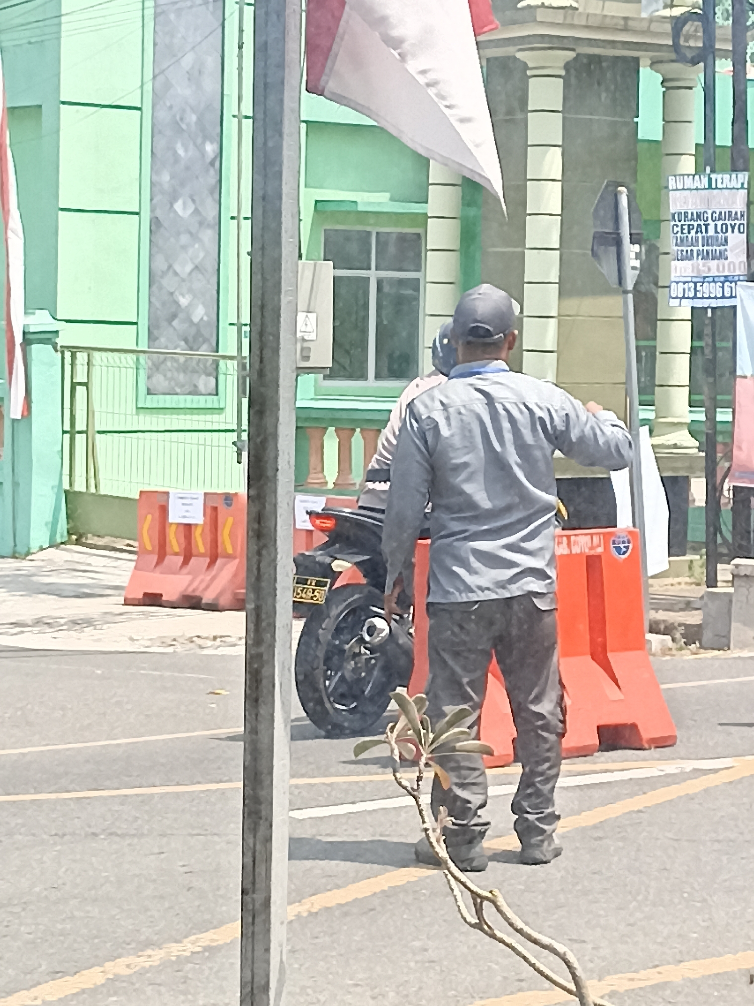 The image depicts a street scene in Pulisen, Indonesia. In the foreground, a person wearing a gray long-sleeved shirt, dark pants, and a cap is standing facing away from the camera. They appear to be gesturing with their right hand. To their left is a motorcycle with a yellow license plate. The motorcycle is partially obscured by a utility pole.
In the background, there are orange traffic barriers with yellow markings. Behind the barriers, a light green building with white trim and columns is visible. A sign with Indonesian text is attached to a pole in front of the building. A flag, possibly the Indonesian flag, is also visible. The overall environment suggests an outdoor urban setting.