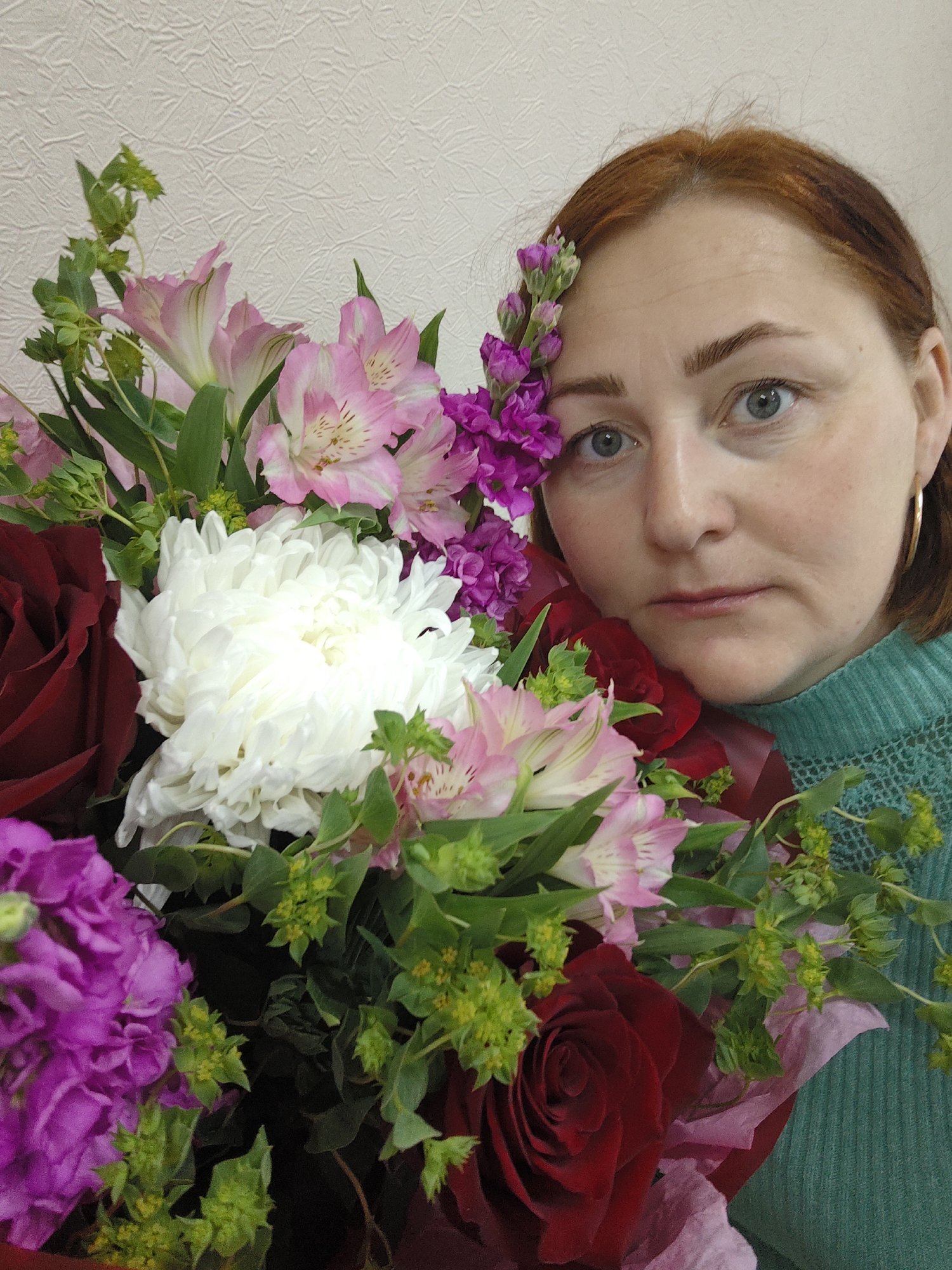 A woman with auburn hair and light blue eyes is holding a large bouquet of flowers. The flowers include dark red roses, white chrysanthemums, pink and white alstroemeria, and purple stock flowers. Some of the stock flowers are placed next to her ear. She is wearing a green sweater and gold hoop earrings. The background is a textured beige wall. Location: Markovskii, Russia.