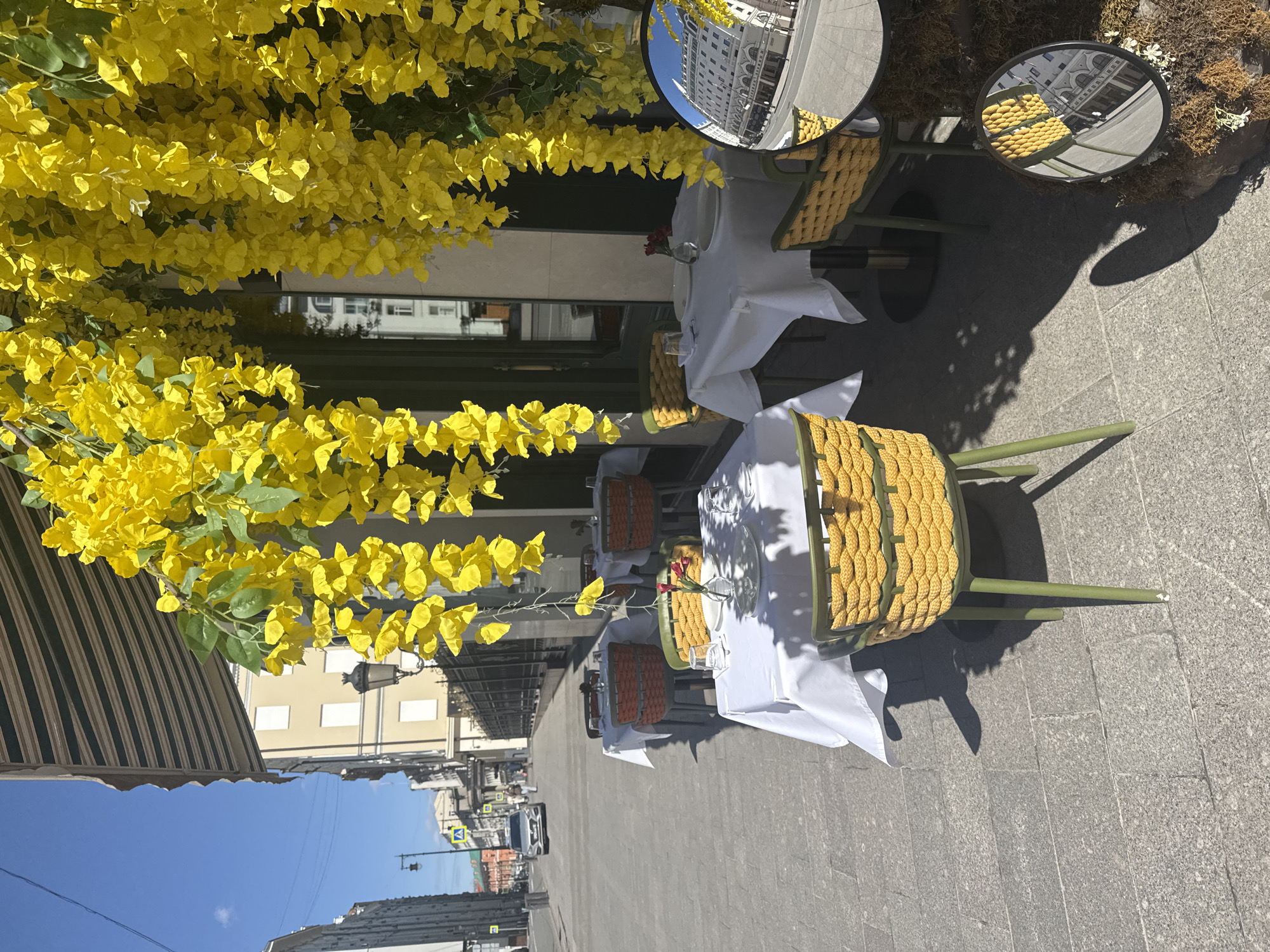 The image shows an outdoor cafe setup on a city street. There are tables covered in white tablecloths, set with glassware and dishes. The chairs are green with yellow woven backs. Two round mirrors are present. Yellow flowers cascade from above, and reflections of buildings are visible in the mirrors. The street extends into the distance with buildings and traffic signals. The image is taken in Moscow, Russia, given the architectural style of the buildings reflected in the mirrors.