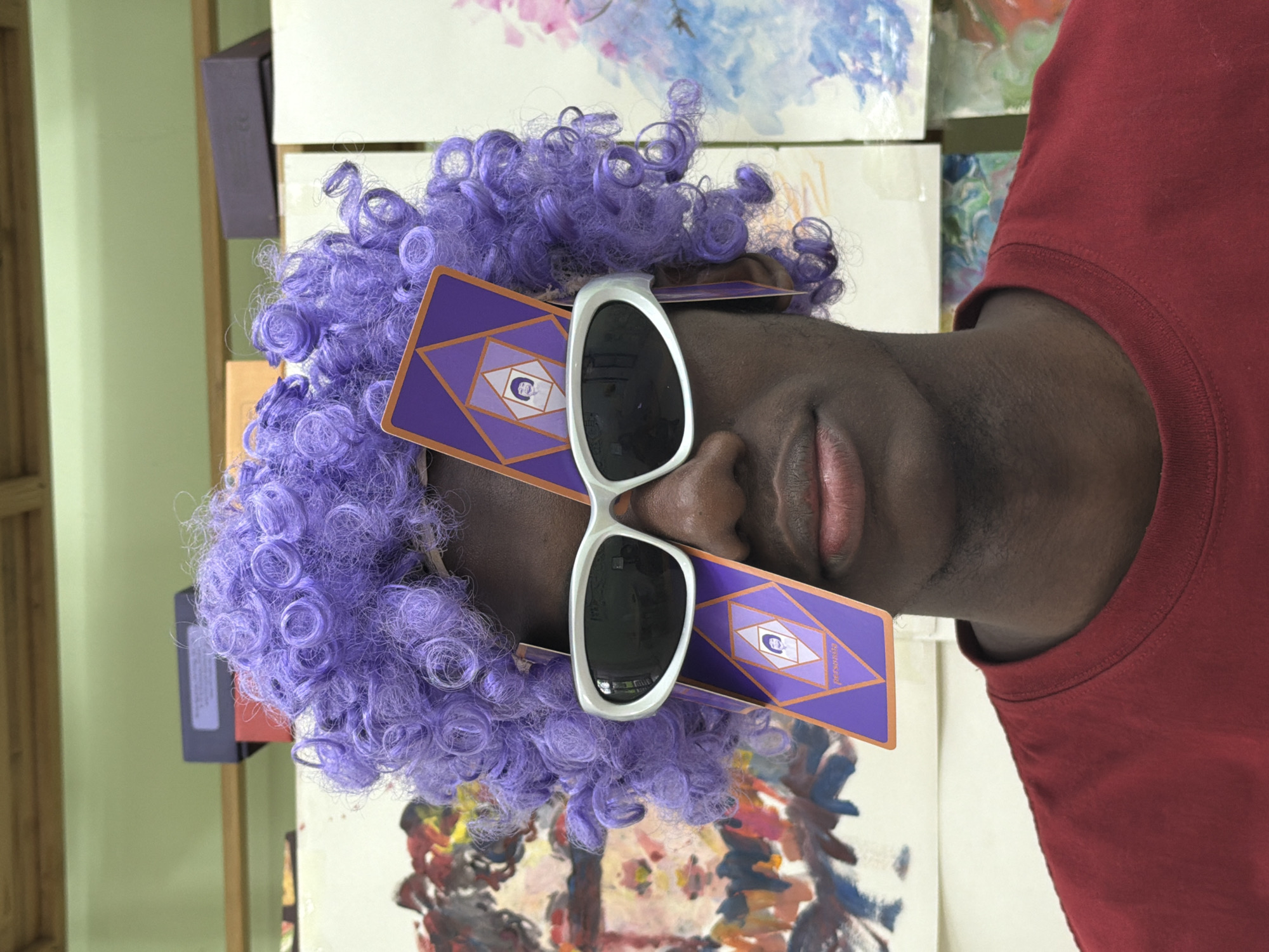 The image shows a person with a purple afro wig wearing large white sunglasses. Two purple and orange cards, possibly tarot cards, are placed over the sunglasses, obscuring the eyes. The person is wearing a red t-shirt. Behind the person, there are several paintings on a wall. The environment appears to be an art studio or a similar creative space. The image does not contain any information that would allow for the identification of its location in the city of Jiangjin, China.