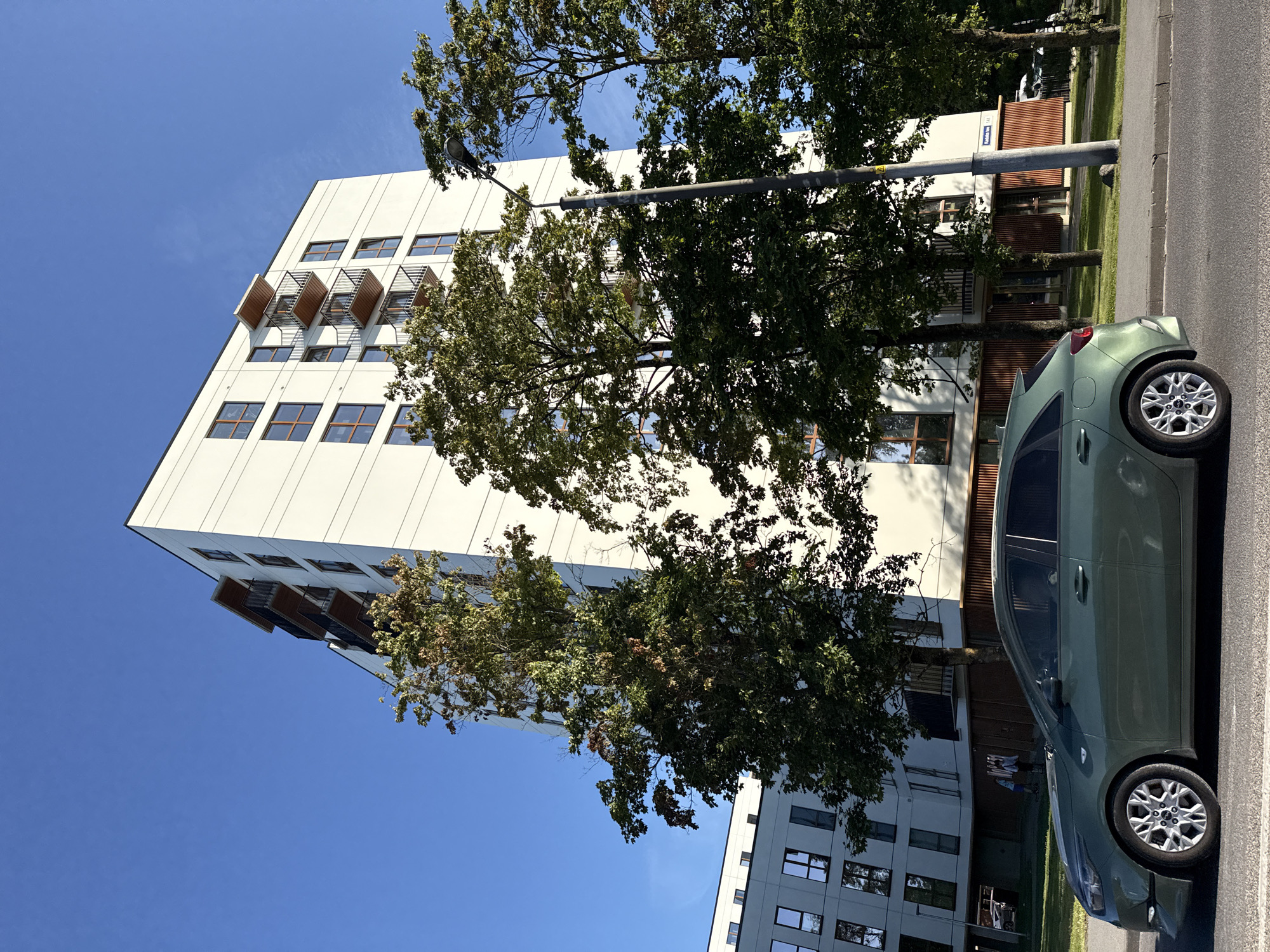 The image depicts a contemporary residential building in Tallinn, Estonia, viewed from street level. A dark green Kia Ceed automobile is parked in the foreground, facing left, on a paved street. The building, a multi-story white structure with brown-trimmed windows and balconies, occupies the middle and background. Several trees with green foliage stand between the car and the building. The sky is clear and blue. A streetlamp is visible to the right of the building. The overall scene suggests a sunny day in a residential area of the city.