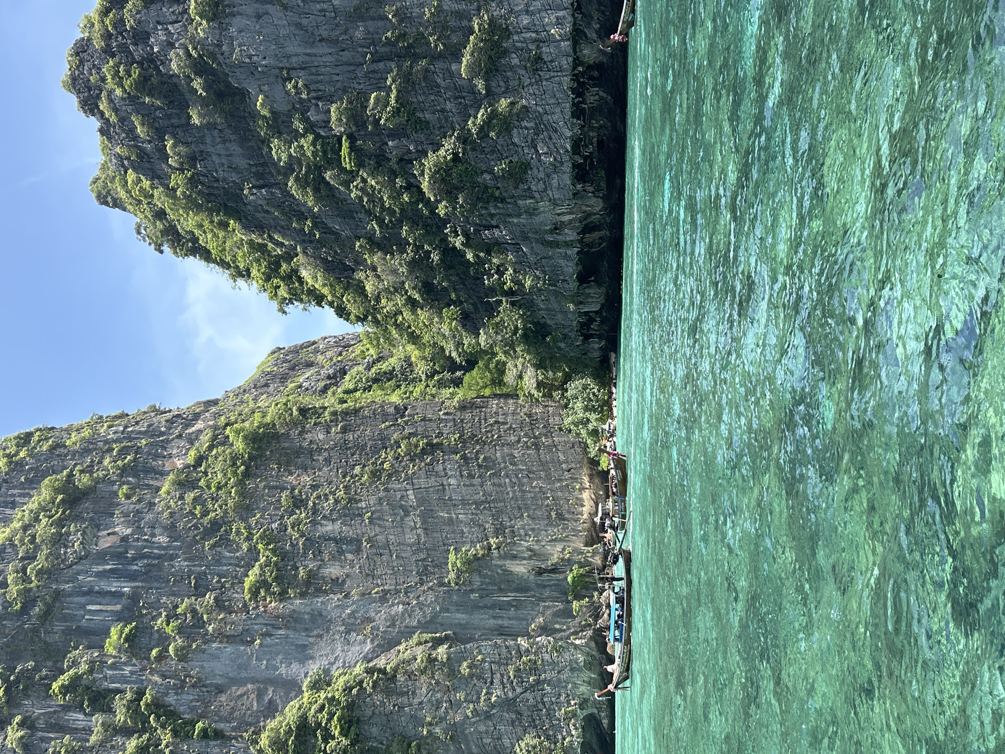 The image depicts a tropical scene in Ao Nang, Thailand. Towering limestone cliffs covered in lush green vegetation rise dramatically from a calm, crystal-clear turquoise sea. Several longtail boats, traditional Thai vessels, are moored in the shallow water near the shore, with a few people visible on board and swimming. The water is so clear that the seabed is visible beneath the surface. The sky above is a clear blue with a few scattered white clouds. The overall impression is one of natural beauty and tranquility, characteristic of the popular tourist destination.