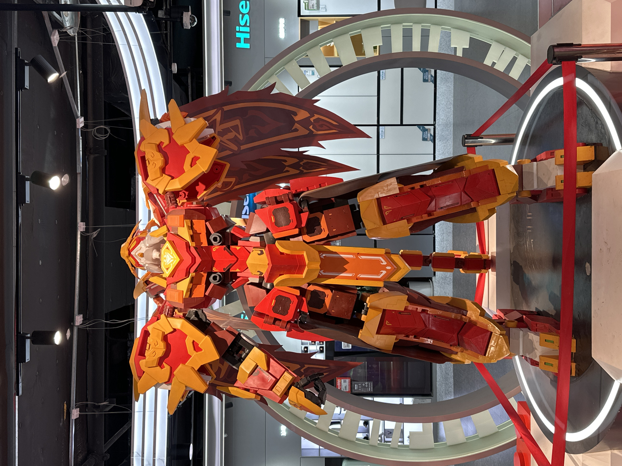 The image shows a large, red and yellow robotic figure, likely a Gundam or similar mecha, displayed in a commercial setting in the Jiulongpo District of China. The robot has a complex, angular design with wing-like appendages and is positioned within a circular, illuminated structure. Red ribbon acts as a barrier, suggesting the display is roped off. The background reveals a commercial environment, possibly a retail store, with fluorescent lighting and other displays. The presence of the Hisense sign indicates a possible product display area. The image appears to be rotated 90 degrees counter-clockwise.