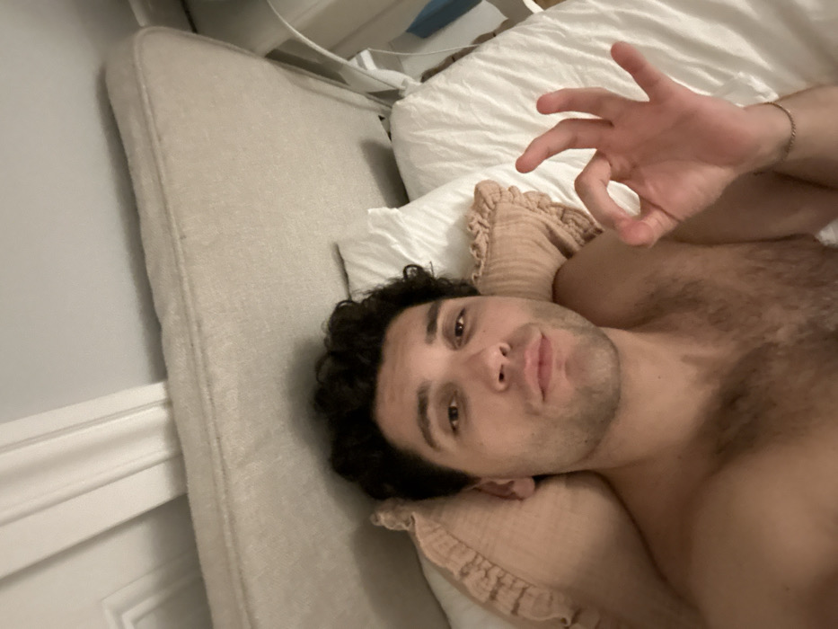 A man lies on his side in bed, his head resting on a ruffled-edge pillow. He has dark, curly hair and stubble. He is shirtless, and some chest hair is visible. His right hand is raised, making an "OK" gesture. White pillows are behind him, and a gray headboard is to his left. A silver chain bracelet is on his right wrist.