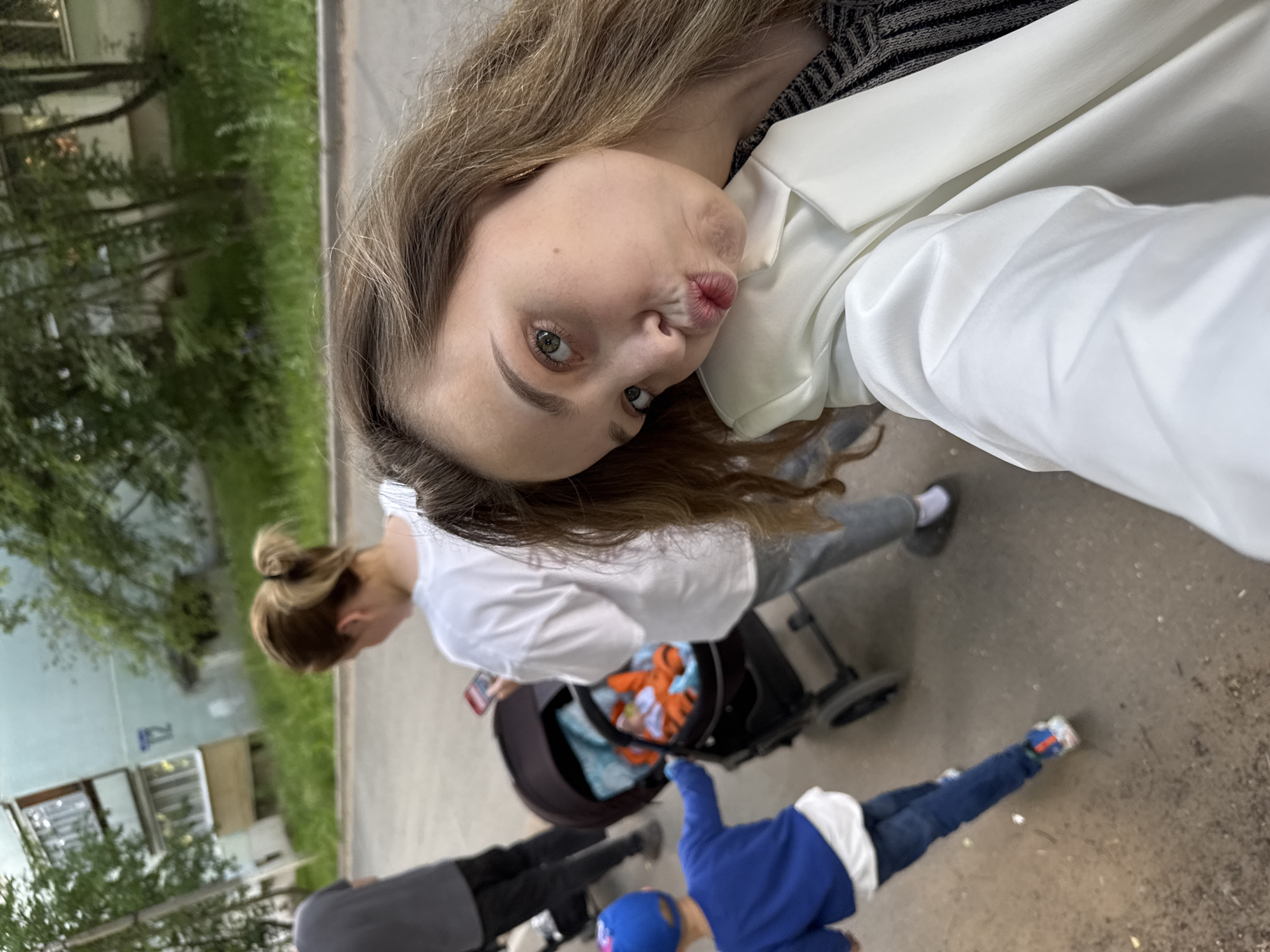This is a vertically oriented, slightly blurry selfie taken outdoors. The primary subject is a young woman in the foreground, looking towards the camera with her lips puckered. She is wearing a white jacket over a striped top. Her hair is long and light brown, falling over her shoulders.
Behind her, a woman with her hair in a bun is pushing a stroller. The stroller appears to contain orange and blue items. Further in the background, a child in a blue jacket and jeans is visible, possibly standing or playing. Another person in dark clothing is partially visible to the left of the child. The setting is a paved area next to a grassy strip and a light blue building with windows. Trees are also visible, suggesting a residential area. The lighting indicates it is daytime, likely overcast. The overall mood seems casual and candid. No visible text is present in the image.