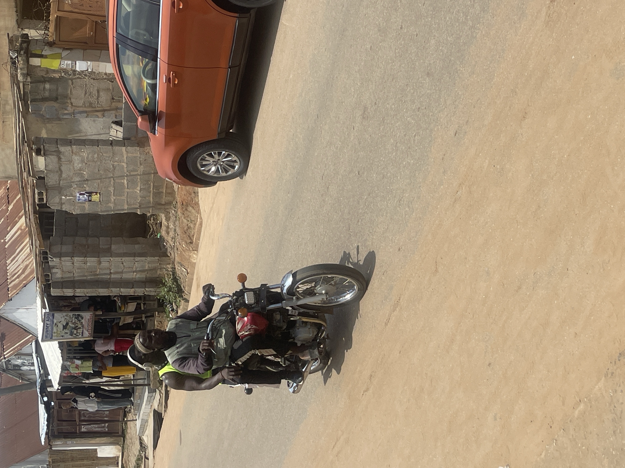In a sunlit outdoor scene in Abuja, Nigeria, a man rides a motorcycle. The man is wearing a green vest over a darker shirt, a white cap, and headphones. He is looking forward as he operates the motorcycle, which is a red and silver model with a passenger seat.
In the background, an orange car is parked. Next to the car is a building constructed with grey concrete blocks, partially covered by a corrugated metal roof. Further to the left, a row of stalls with similar corrugated metal roofs is visible, indicating a market or commercial area. Some of these stalls display goods.
One stall has a sign that reads "MUMMY ADA" and another smaller sign with a phone number "081 3592694". The overall impression is a busy street scene during daylight. The weather appears to be sunny, casting shadows on the ground. The scene depicts everyday life in a Nigerian urban environment.