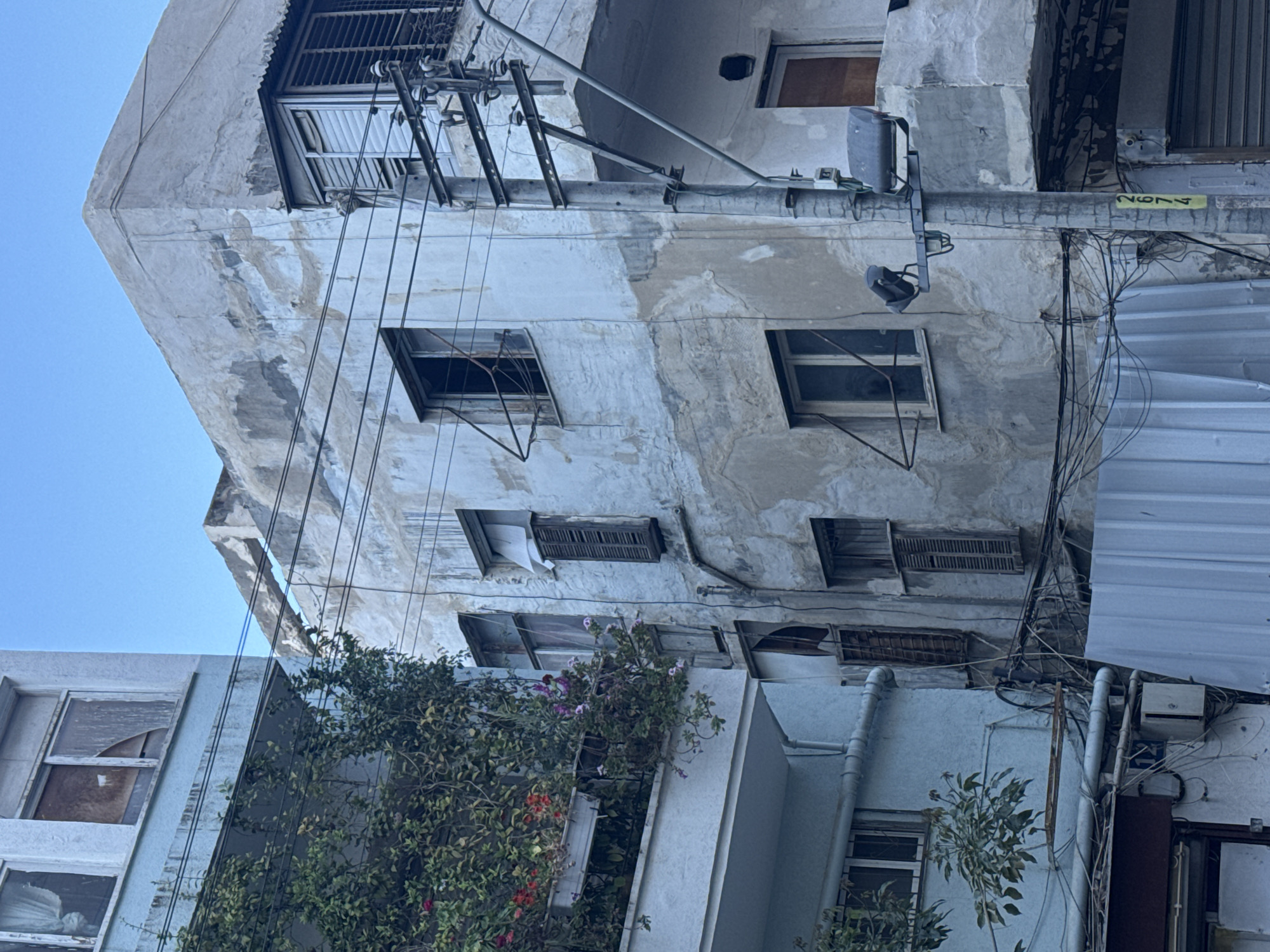 The image depicts a densely populated urban scene, presumably in Tel Aviv, Israel, given the architecture and style of the buildings. The focus is on a dilapidated building with visible signs of wear and tear on its facade, including peeling paint, water stains, and aging concrete. The building has multiple windows, some of which are covered with shutters or grills. A tangle of electrical wires and utility poles are visible, adding to the dense urban atmosphere. A section of corrugated metal siding is also visible, possibly used as a makeshift repair. Below the weathered building, greenery in the form of vines and shrubs adds a contrast to the harsh urban environment.
