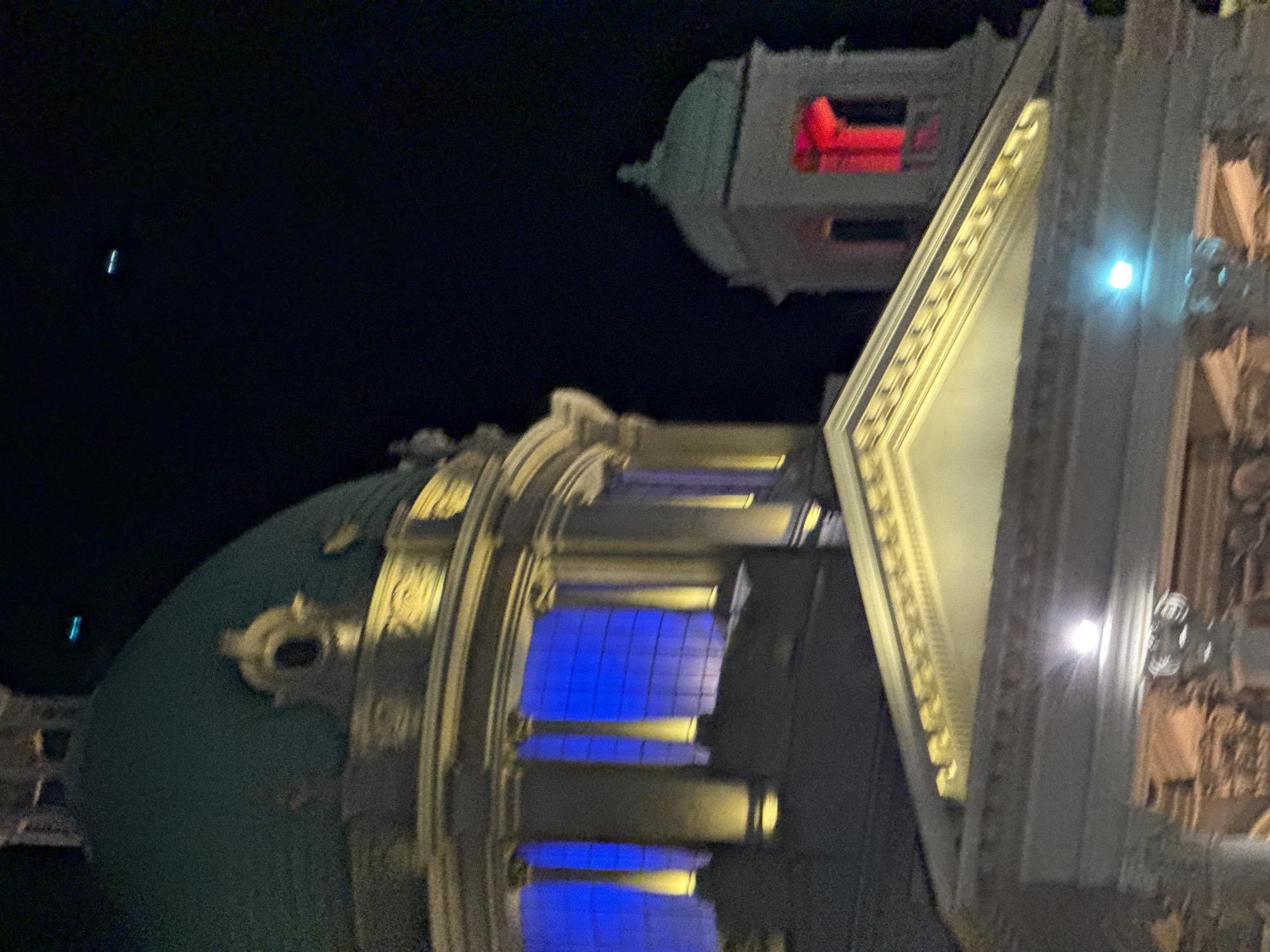 At night, a low-angle shot captures an ornate building illuminated with colored lights. A green dome structure is on the left, adorned with decorative elements. Windows are illuminated in blue. Adjacent to the dome is a section of the building with gold trim and detailed carvings, reflecting artificial light. In the upper portion of the frame, a smaller turret-like structure is lit with red light. The backdrop is a dark night sky. It is the National Assembly Building in Belgrade, Serbia.