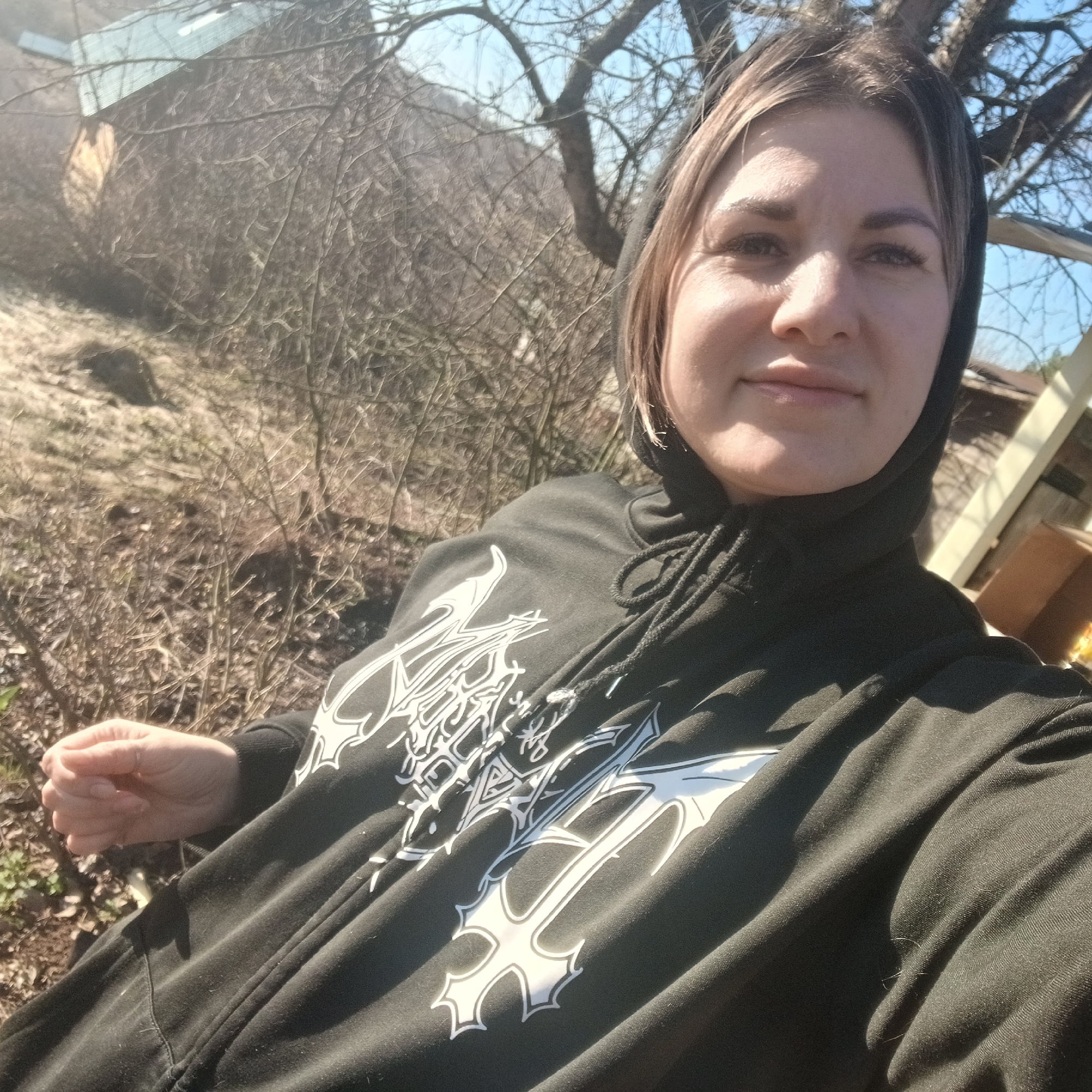 A person is outdoors on a sunny day. The person is wearing a black hooded sweatshirt with a white graphic print on the front. The background includes bare trees, a hillside, and a building. The location is Unknown, Unknown.