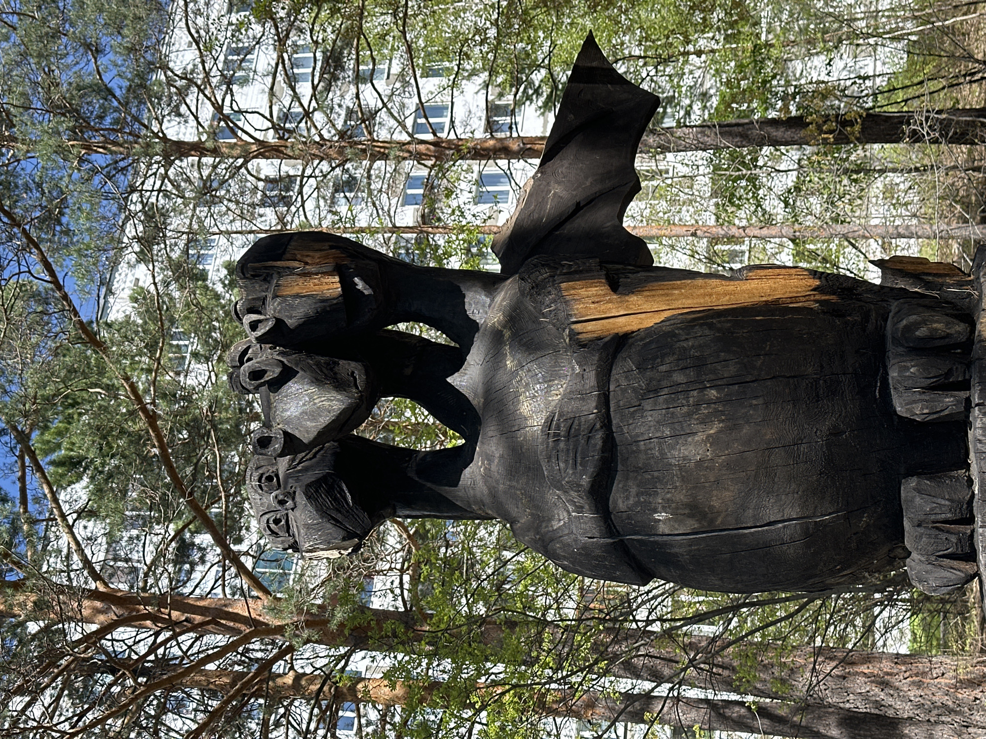 The image depicts a large, dark, wooden sculpture in an outdoor setting. The sculpture appears to be of a dragon or mythical creature, with visible wings, a stout body, and multiple faces with prominent eye holes. The wood has a charred or burned appearance, adding to its unique texture and aesthetic.
The sculpture is positioned in front of a backdrop of trees and a building with multiple windows, suggesting an urban park or landscaped area. Based on the location in Sayansk, Russia, it's likely situated in a public space like a park or recreational area. The environment includes both deciduous and coniferous trees, indicating a temperate climate. The branches and leaves suggest it is spring or summer. The presence of a building with modern architecture suggests the sculpture is located within or near a residential or commercial area of the city.