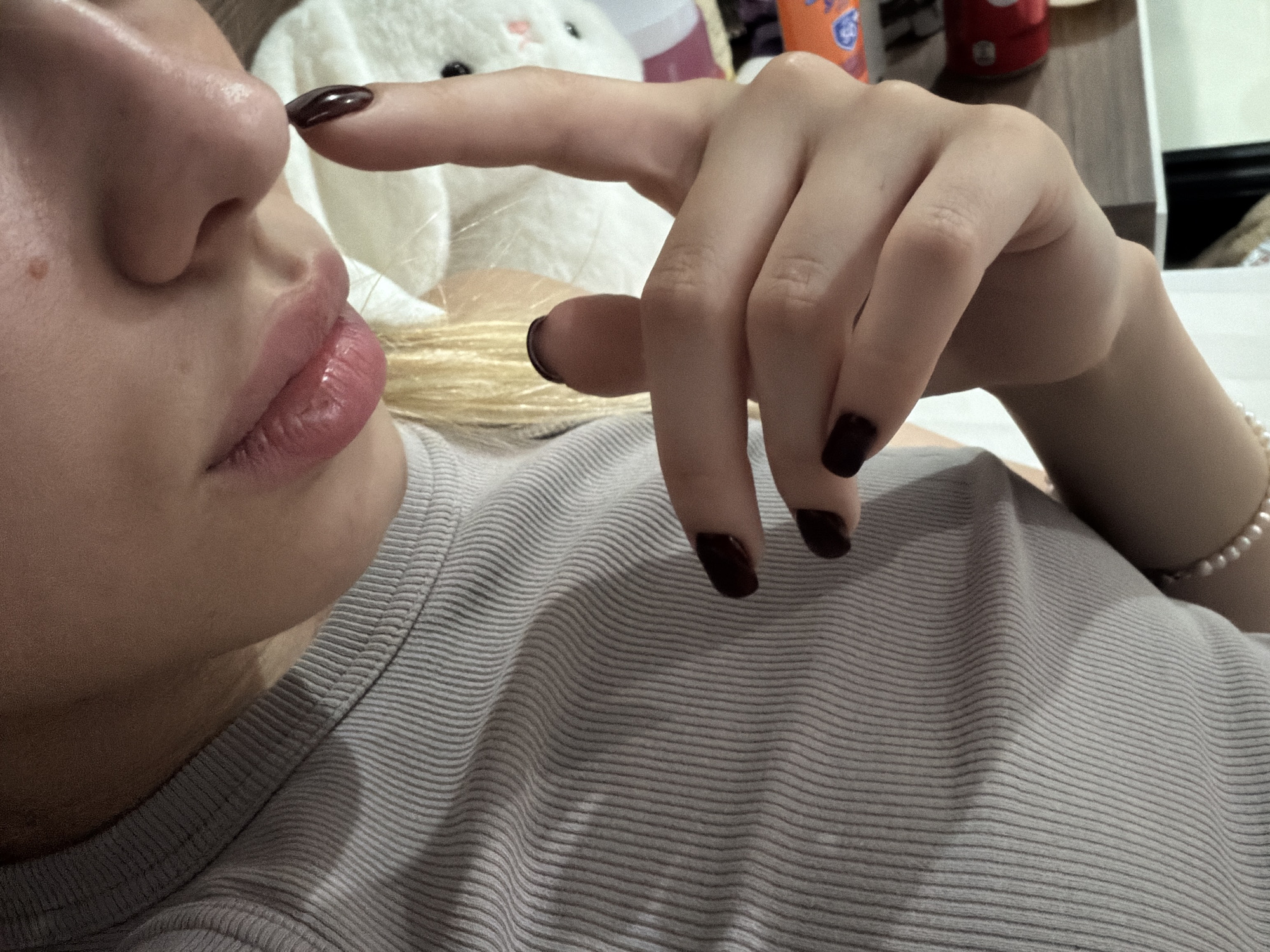 The image shows a close-up of a person's face and upper body, with a focus on their lips, nose, and hand. The person has fair skin, pink lips, and blonde hair that is partially visible. Their fingernails are painted a dark, glossy color. The person is wearing a light gray, ribbed t-shirt. The background is blurred but appears to be indoors, with a white stuffed animal and some objects on a shelf visible. There is no indication of the city of Đà Nẵng, Vietnam, in the image.