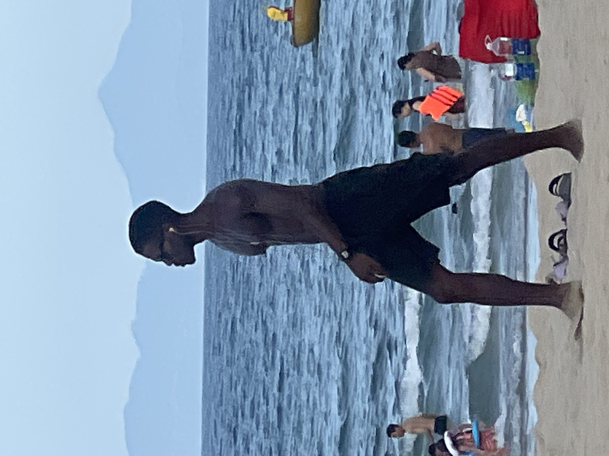 The image shows a person of African descent walking on a sandy beach in Đà Nẵng, Vietnam. The person is wearing dark shorts and sunglasses and has earbuds in their ears. In the background, there are other beachgoers in the ocean, and a yellow boat with a person in it is visible on the water. The ocean is blue and has small waves. In the far distance, mountains are visible against a pale blue sky. On the sand in the foreground, there are a pair of flip-flops and a red bag with water bottles.