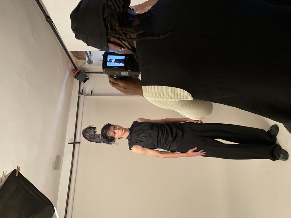 A photographer captures a fashion model in a studio setting. The model, wearing a dark outfit and feathered headpiece, stands against a white backdrop. The photographer, clad in black with dreadlocks, holds a camera. Lighting equipment is visible in the background, suggesting a professional photoshoot.