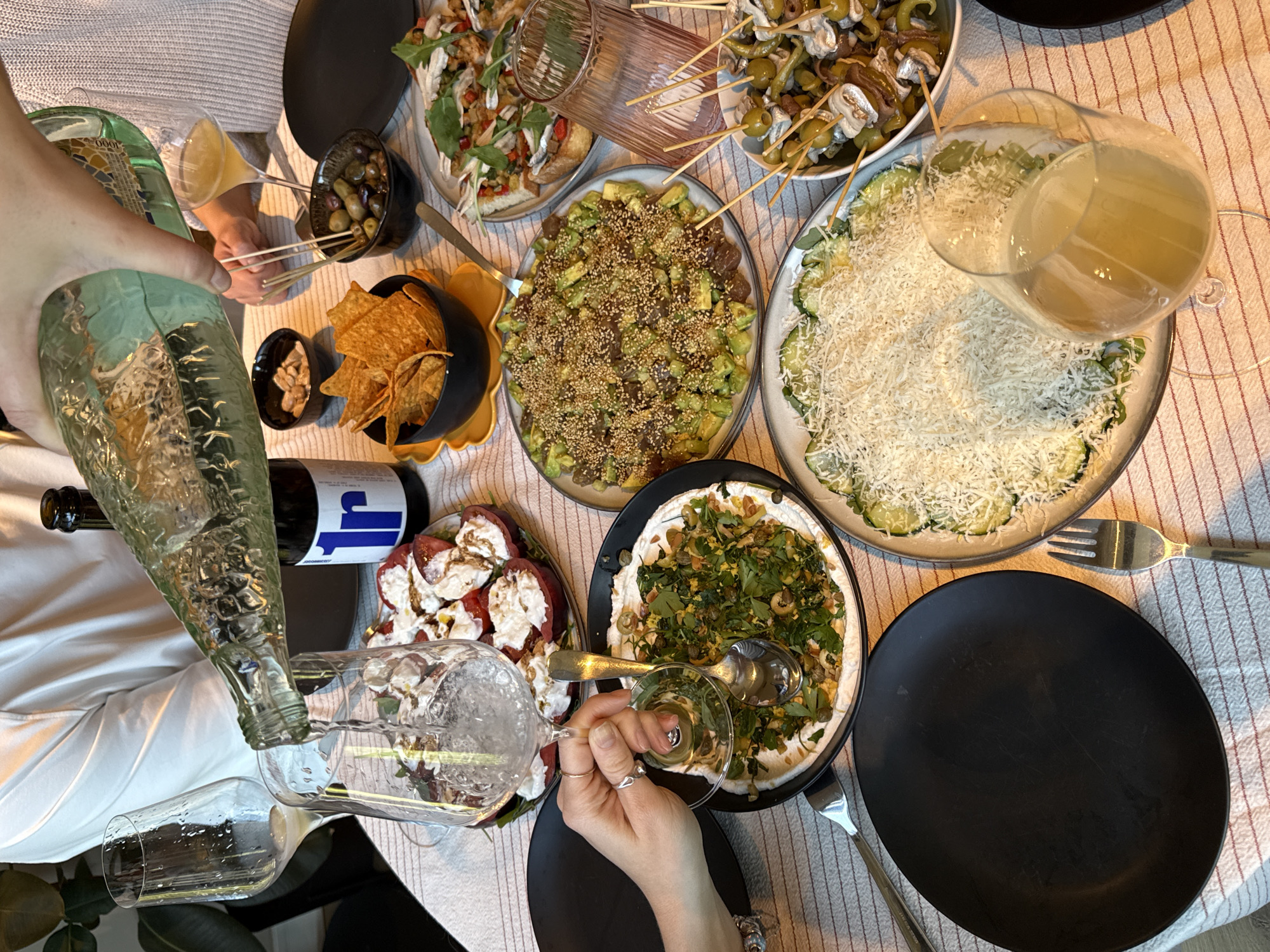 A top-down view reveals a table laden with diverse foods and beverages, suggesting a gathering or meal in progress. The table is covered with a light-colored tablecloth featuring subtle striped details. The dishes presented include a pizza with tomatoes, fresh basil, and other unidentified toppings; another dish with a mixture of avocado and seeds; a zucchini salad topped with shredded cheese; a plate filled with caprese salad consisting of tomatoes and mozzarella; a bowl of tortilla chips alongside a bowl of olives, and skewers with olives, anchovies, and peppers. A green bottle is being poured into a glass, with another glass and bottle nearby. The scene is in Barcelona, Spain.