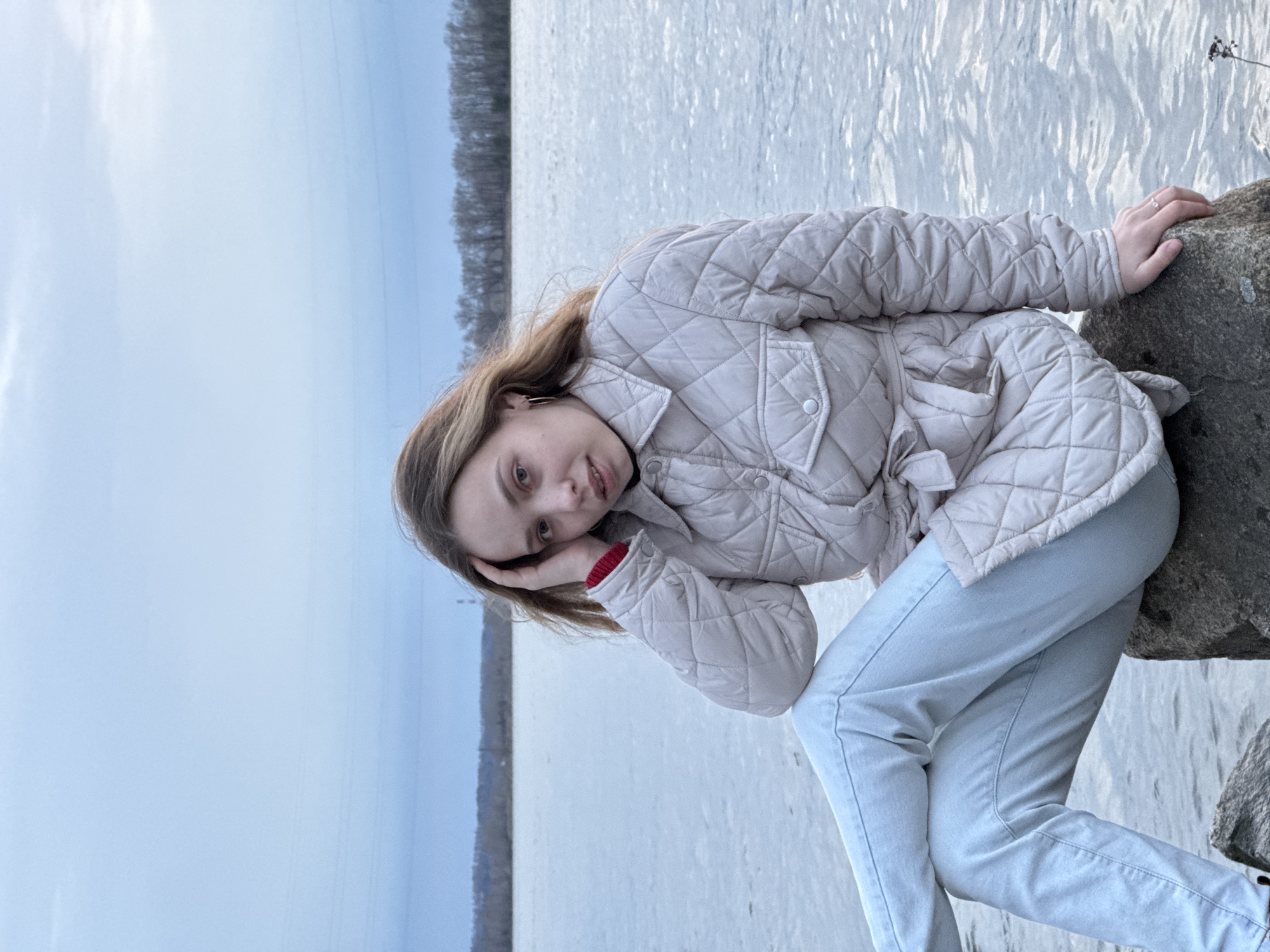 A young woman is prominently featured in this outdoor portrait, positioned by a large body of water in Chaikovsky, Russia. She has light brown hair and is wearing a light, possibly cream-colored, quilted jacket with a collar and pockets, and light blue jeans. A red cuff is visible from an undershirt on her left arm. She wears a ring on the fourth finger of her left hand.
The woman is looking directly at the camera with a gentle, soft smile and a relaxed expression. Her head is tilted slightly to her right, and her left hand is casually placed near her head. She is sitting or leaning on a large, grey rock that occupies the right side of the frame.
Behind her, a vast expanse of water, likely a lake or river, stretches across the middle ground, its surface appearing calm with subtle ripples reflecting the light. In the far background, a dense line of bare trees forms a dark, low horizon against a pale, overcast sky. The lighting suggests an overcast day, contributing to the soft, diffused illumination of the scene. There is no visible text in the image.