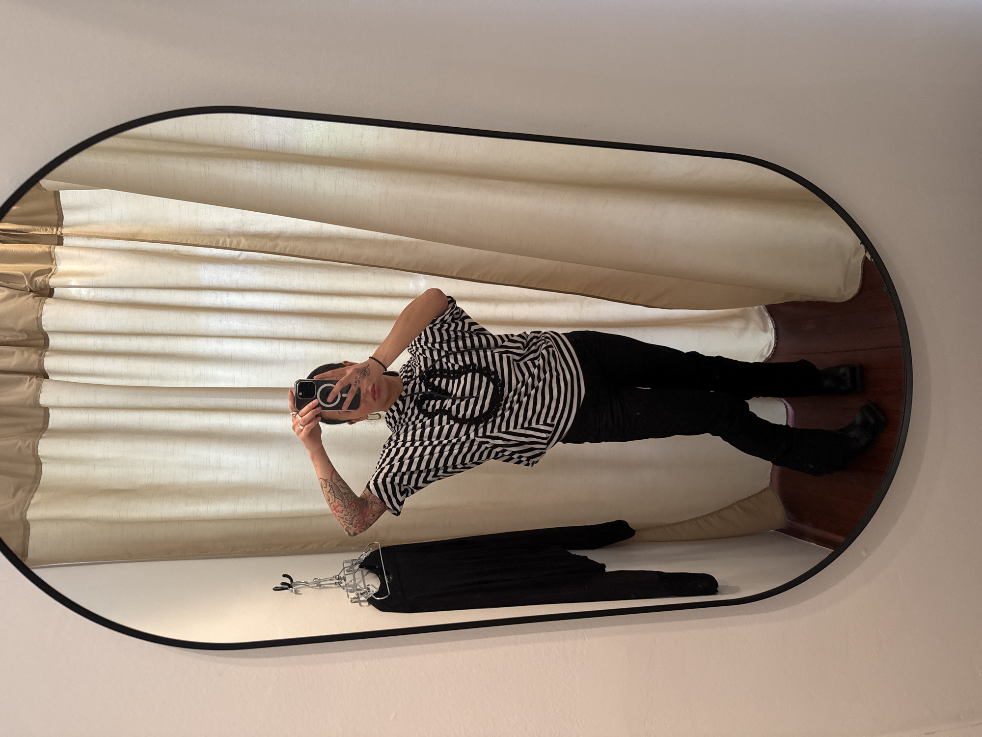 A person is taking a selfie in a mirror. The mirror is oval with a black frame. The person is wearing a black and white striped shirt and black pants. They have tattoos on their arm. A black garment hangs on a hanger in the foreground of the mirror's reflection. Beige curtains are in the background. The location cannot be determined from this image.