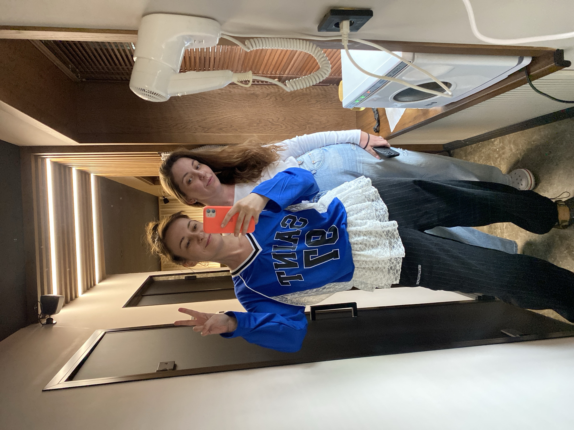 Two women are posing for a selfie in what appears to be a bathroom. One woman is holding the phone, wearing a blue jersey-style top with white lace and black striped pants, and holding up a peace sign. The other woman is behind her, wearing a white long-sleeved shirt and jeans, holding a phone in her hand. A hair dryer and a bidet are mounted on the wall. The bathroom has a wooden accent wall and light fixtures. The specific location in Votkinsk, Russia, cannot be determined from this image.