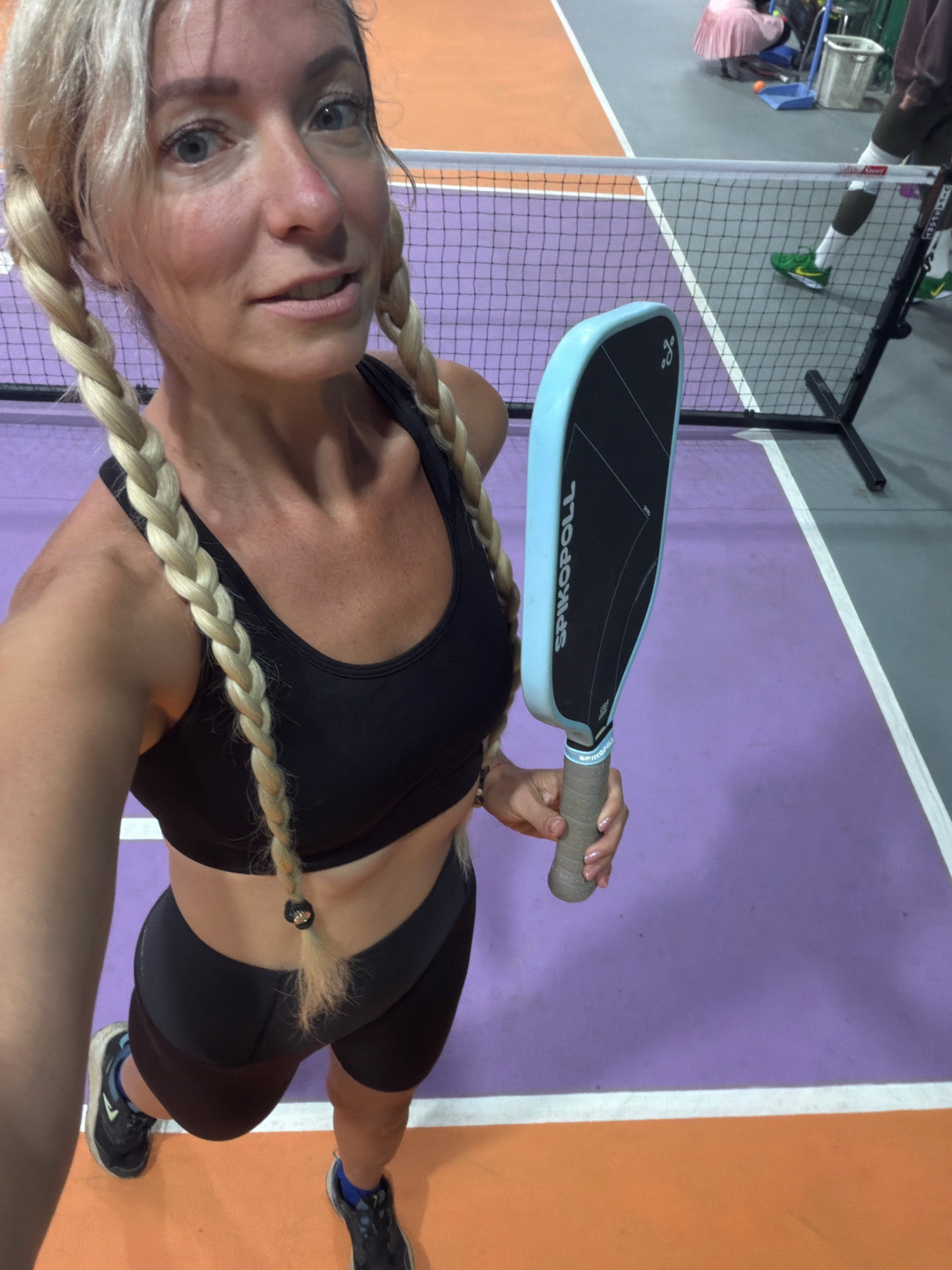 The image shows a woman in athletic wear holding a pickleball paddle. She has blonde hair in two braids and is standing on a pickleball court. The court is marked with purple and orange lines. In the background, a net and other players can be seen. The woman is smiling and looking at the camera. The scene appears to be indoors. The location in the city of Da Lat, Vietnam is not discernible from the provided image.