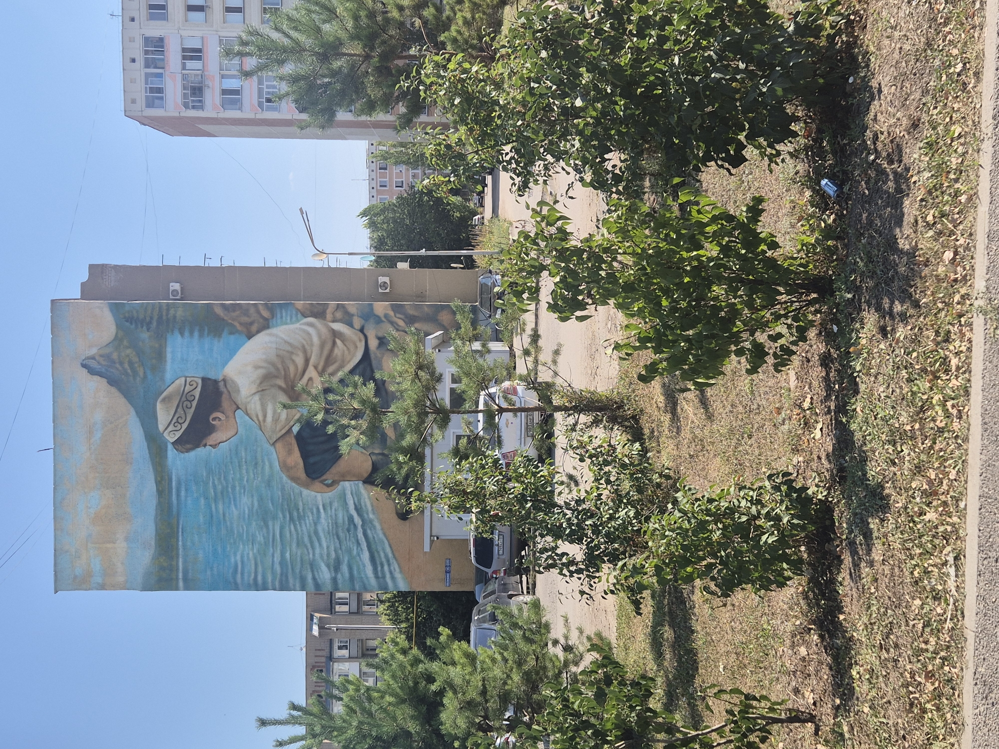 A large mural on the side of a multi-story apartment building depicts a young boy wearing a traditional hat and looking out at a body of water with mountains in the background. The building is concrete and light-colored, with several air conditioning units visible on its facade. To the right of this building, another similar apartment building is visible. In the foreground, there are several evergreen trees and some smaller leafy bushes. The ground is covered with dry grass and fallen leaves. A paved road runs in front of the buildings, and several cars are parked along it. The sky is clear and blue. The mural is located on Tchaikovsky Street in Kokshetau, Kazakhstan.