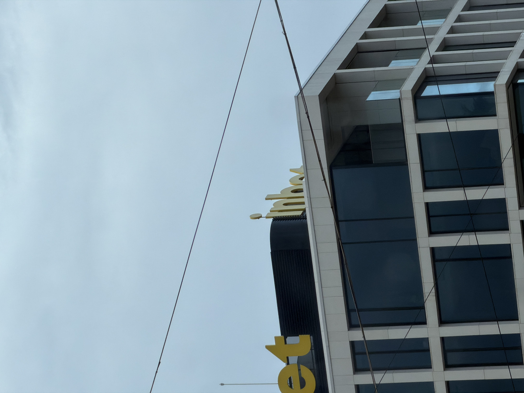 The image shows the side of a modern, high-rise building with a grid-like facade in Sofia, Bulgaria. The building has large, dark windows set into a light-colored frame. A portion of a gold-colored sign, partially obscured, is visible on the adjacent building. Overhead wires stretch across the sky, contrasting with the building's architecture and the overcast sky. The overall view is angled upwards.
