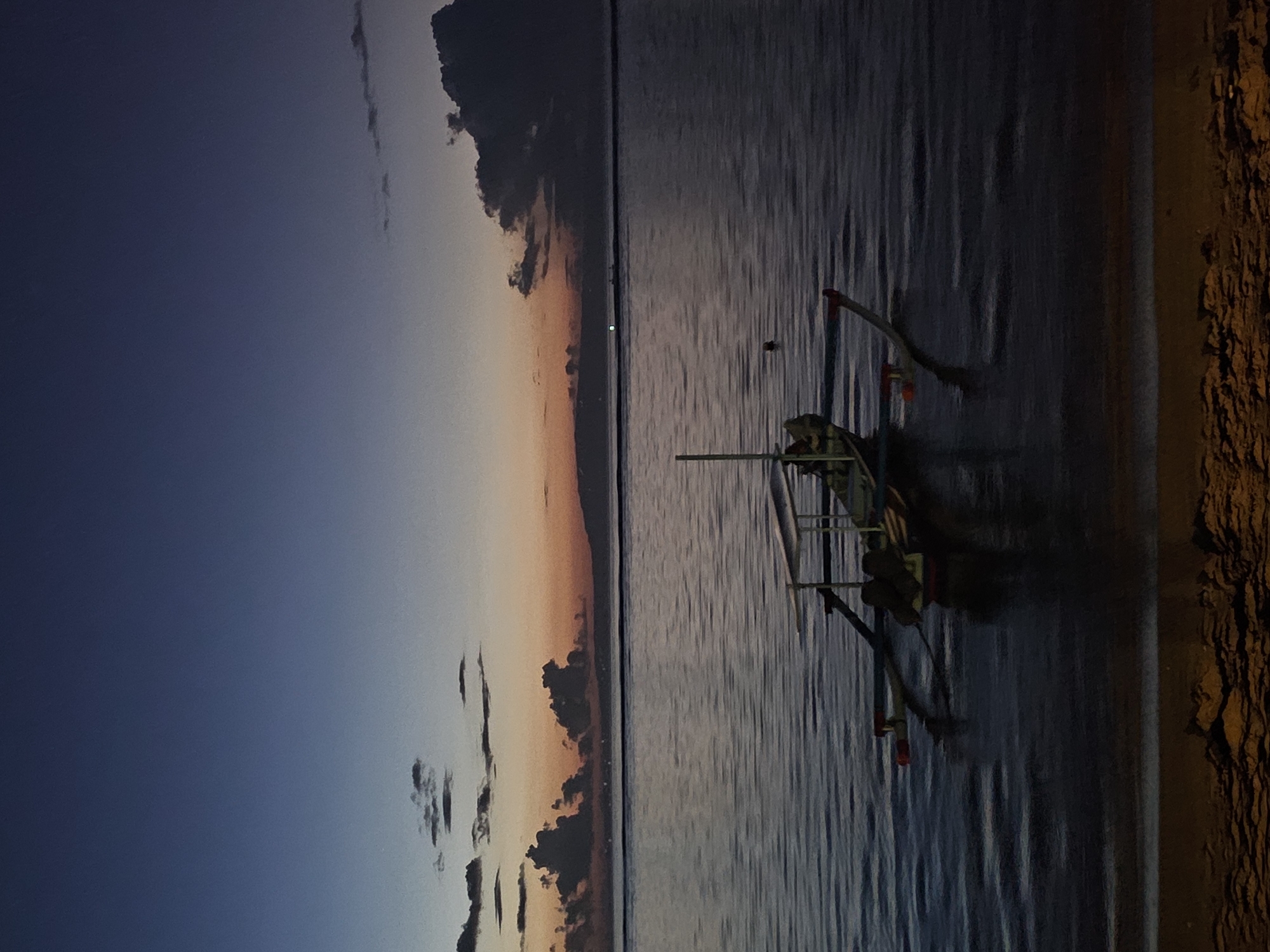 The image shows a traditional outrigger boat on the water, likely in the coastal area of Denpasar, Indonesia. The sky is a mix of blue and orange hues, indicating either sunrise or sunset. The water is relatively calm with subtle ripples. The boat is docked close to the shore, as seen by the rocky texture on the right edge of the image. There are no visible people in the frame.