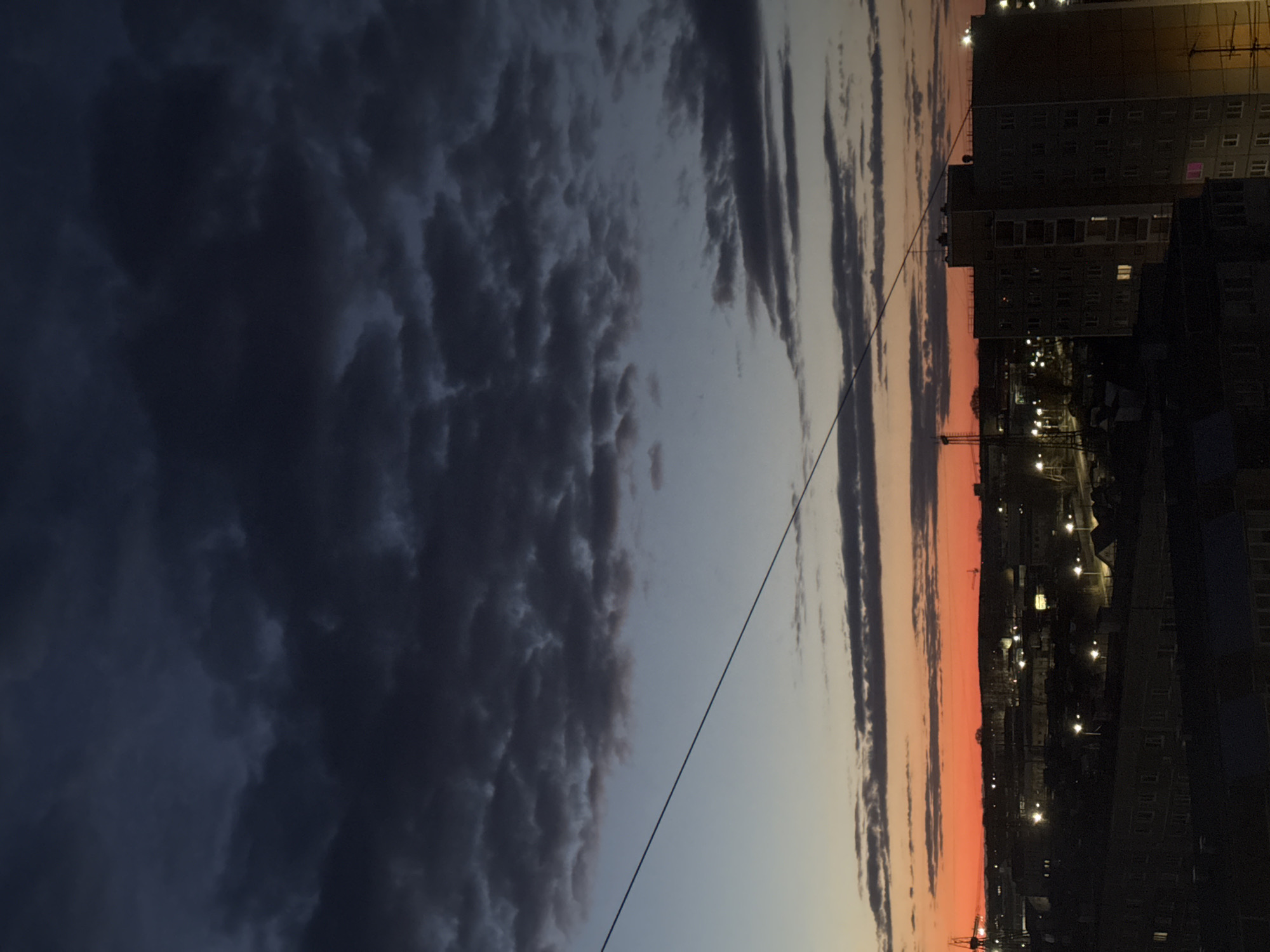 The image displays a dramatic evening sky over the city of Chaikovsky, Russia, characterized by a stark contrast between dark clouds and a vibrant sunset.
The upper-left portion of the frame is dominated by voluminous, dark grey and blue-black clouds, appearing heavy and textured, suggesting overcast or potentially stormy weather. These dense cloud formations extend across a significant part of the sky.
Towards the lower-right, the sky transitions into a brilliant sunset, featuring a striking band of intense orange and pink hues along the horizon. This colorful glow gradually fades into lighter blues and greys higher up, where a few elongated, wispy clouds are silhouetted against the bright sky.
A thin, dark power line or utility cable stretches diagonally across the image, from the upper-left corner towards the lower-right, bisecting the sky.
Below the colorful horizon lies the city's silhouette. Multi-story buildings, appearing to be residential blocks, are visible in dark tones, with some windows illuminated, indicating activity within. Numerous streetlights and other urban lights twinkle across the cityscape, creating bright points of light against the darkened ground and structures. A construction crane is faintly visible against the sunset glow, hinting at urban development.
The scene captures the tranquil yet visually striking transition from dusk to night in an urban environment, emphasizing the dramatic natural light display above the bustling city. No people or visible text are present in the image.