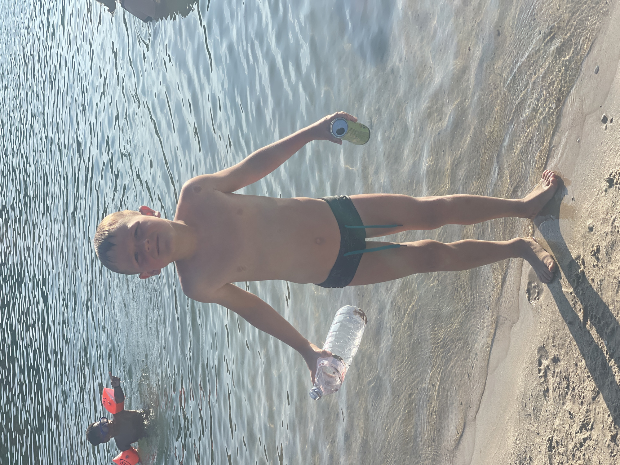 A young boy stands on a sandy shore at the edge of calm, clear water. He is wearing dark green swim trunks. His skin is tanned, and his short hair is light brown. He is holding a clear plastic water bottle in his left hand and a green aluminum can in his right hand. The water reflects sunlight, creating ripples. In the background, another child wearing orange arm floaties and goggles is in the water, pointing. The location is a beach with shallow water.