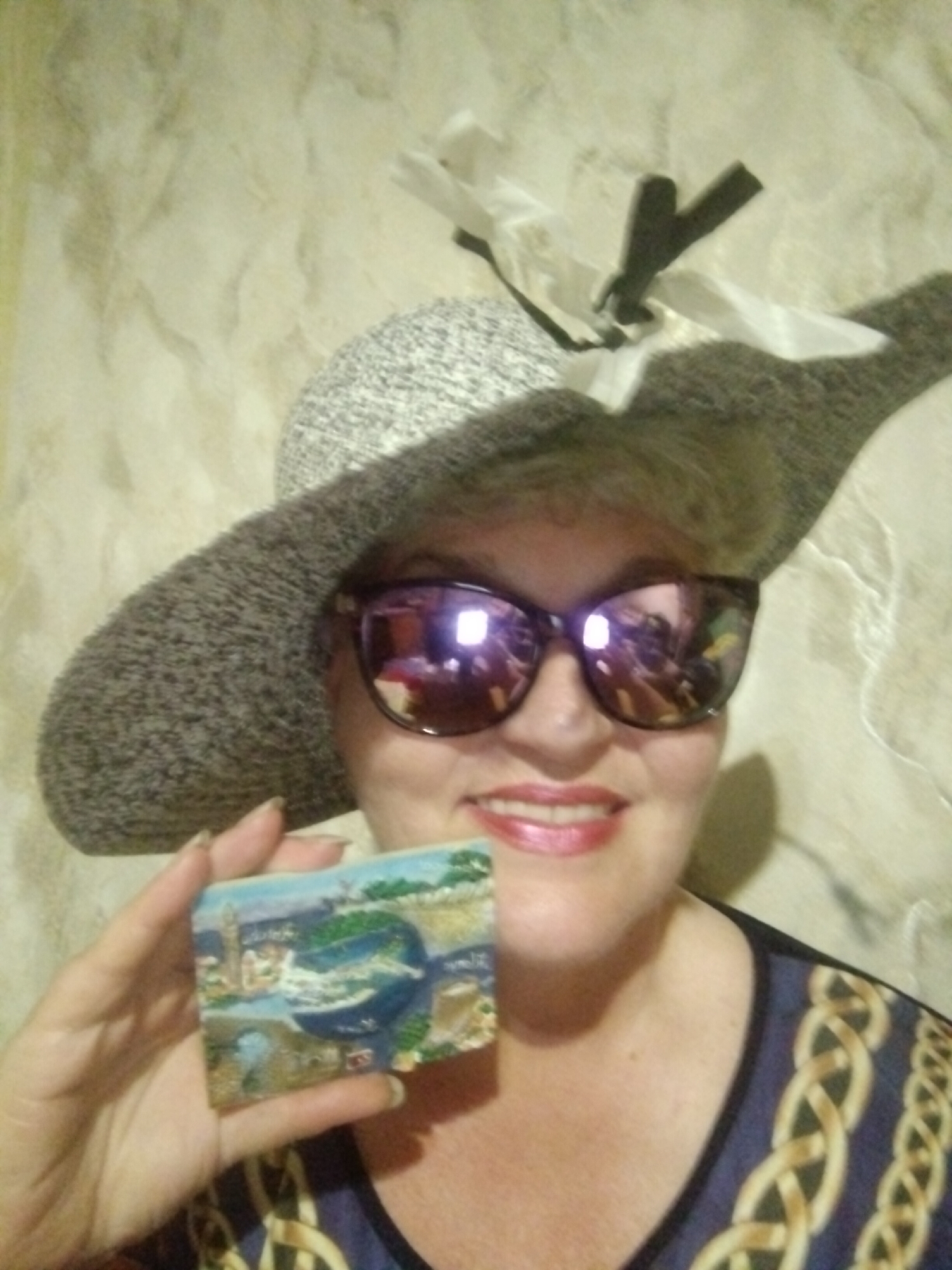 A woman wearing a wide-brimmed hat and sunglasses is holding up a small rectangular magnet. The magnet depicts a scenic view of a city and body of water, possibly a historical or tourist location. The woman's face is partially visible, with her lips painted in a pinkish-red shade. She is wearing a dark top with a gold chain-like pattern. The background appears to be a textured wall. There is no information in the image to suggest the location is in the city of Horishni Plavni, Ukraine.