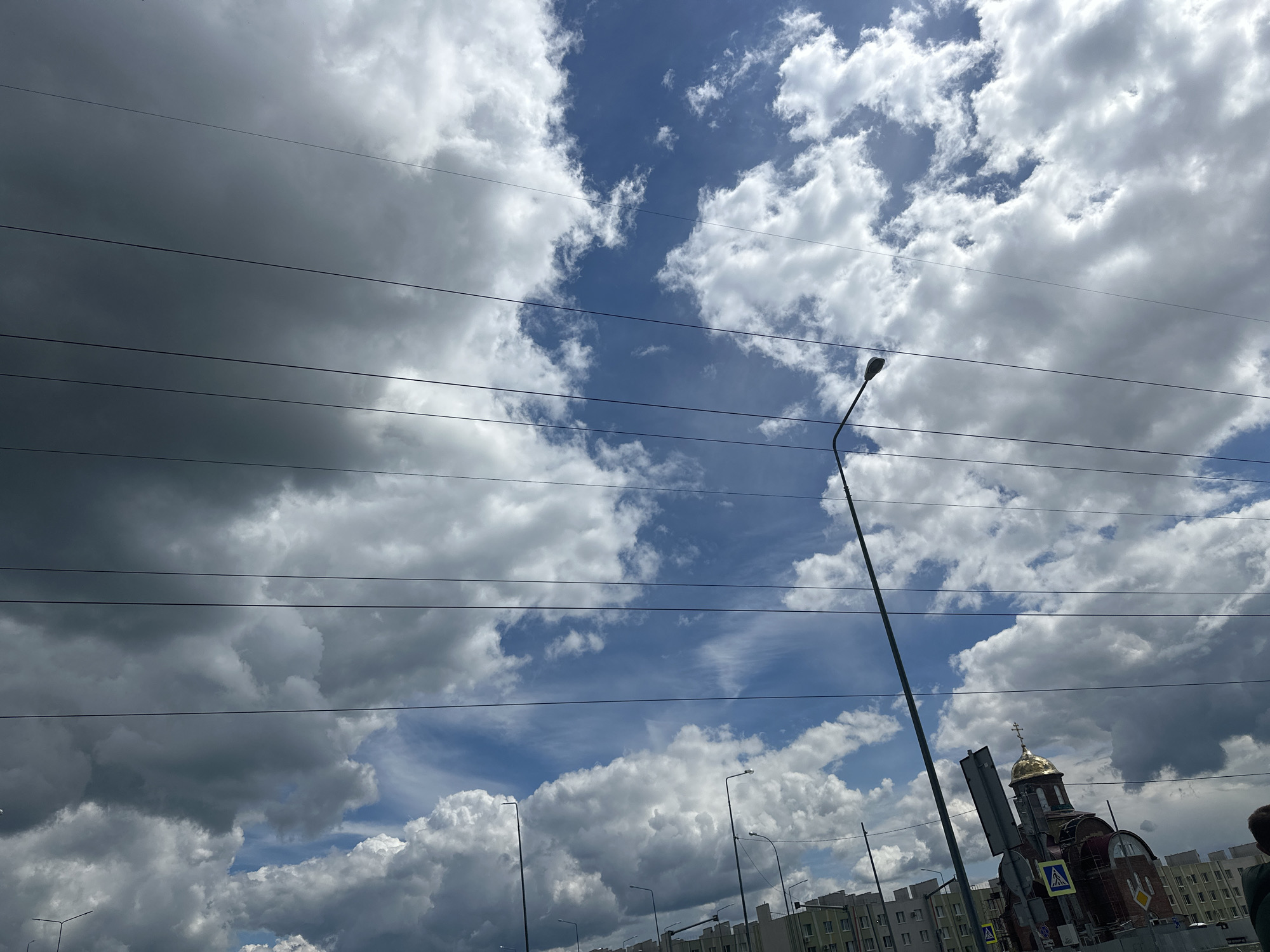 This image, taken in Stroykeramika, Russia, captures a cloudy sky with a mix of dark and light clouds. Several power lines are visible in the foreground, stretching horizontally across the frame. A tall street lamp stands prominently on the right side of the image. In the background, a building with a golden dome, resembling a church, is visible. Several other buildings and street lamps are also present. The overall scene appears to be an outdoor urban environment under a dynamic sky.