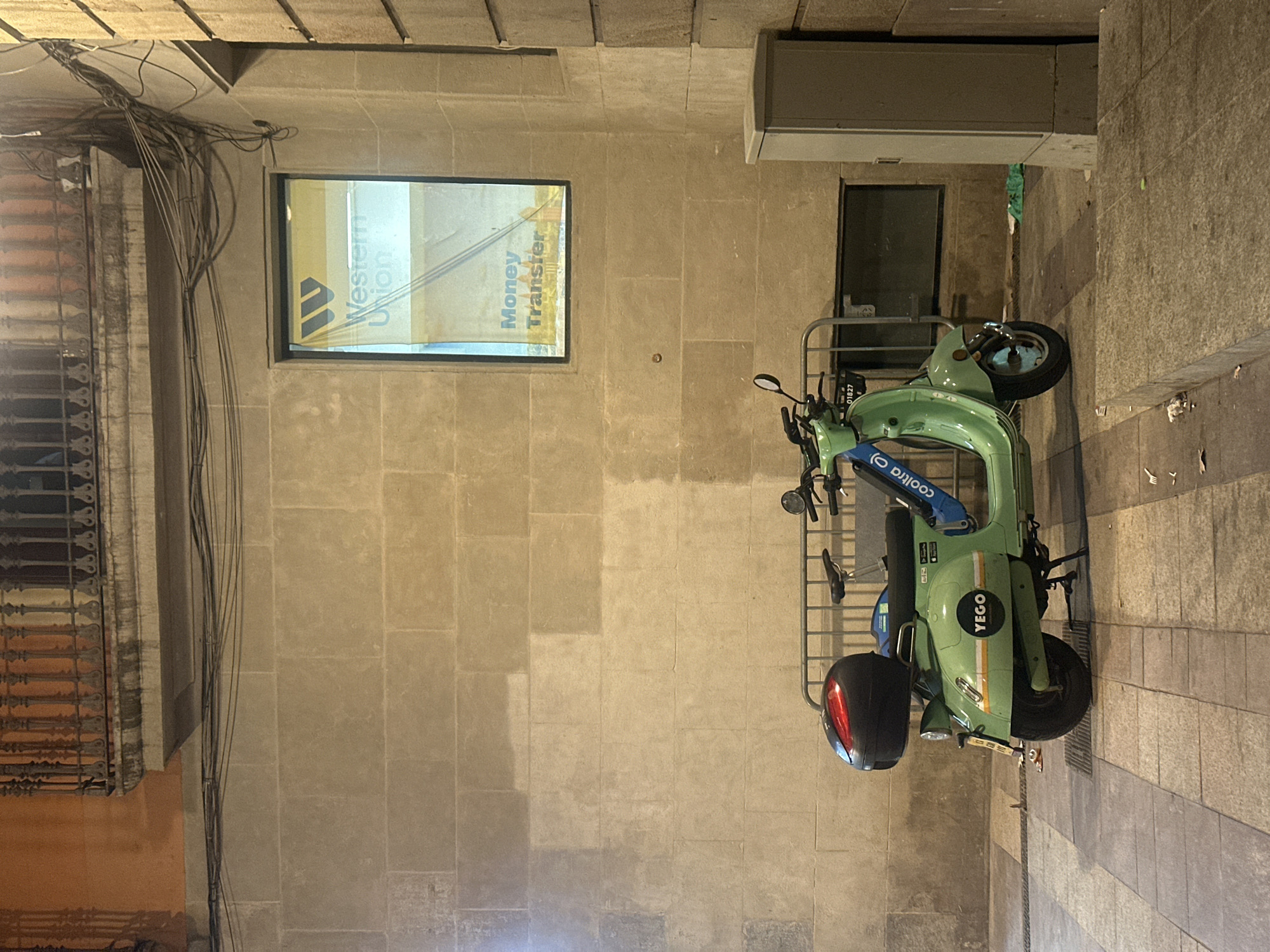 The image depicts a street scene in Barcelona, Spain, at night. The primary subjects are a light green electric scooter and a building facade. The scooter, branded "YEGO," is parked on a cobblestone sidewalk. It is equipped with a black helmet securely attached to the rear. The building facade is constructed of large, light-colored stone blocks and features a window displaying advertisements for "Western Union" and "Money Transfer." Directly above the window, a cluster of black electrical wires hangs precariously. To the right of the scooter, a metal railing encloses a walkway, and a grey utility box is mounted on the building. The overall lighting suggests it is nighttime, with artificial lights illuminating the scene. The atmosphere is calm and quiet, with no people visible. The location cues, such as the scooter's branding and the architectural style, firmly place the scene in Barcelona.