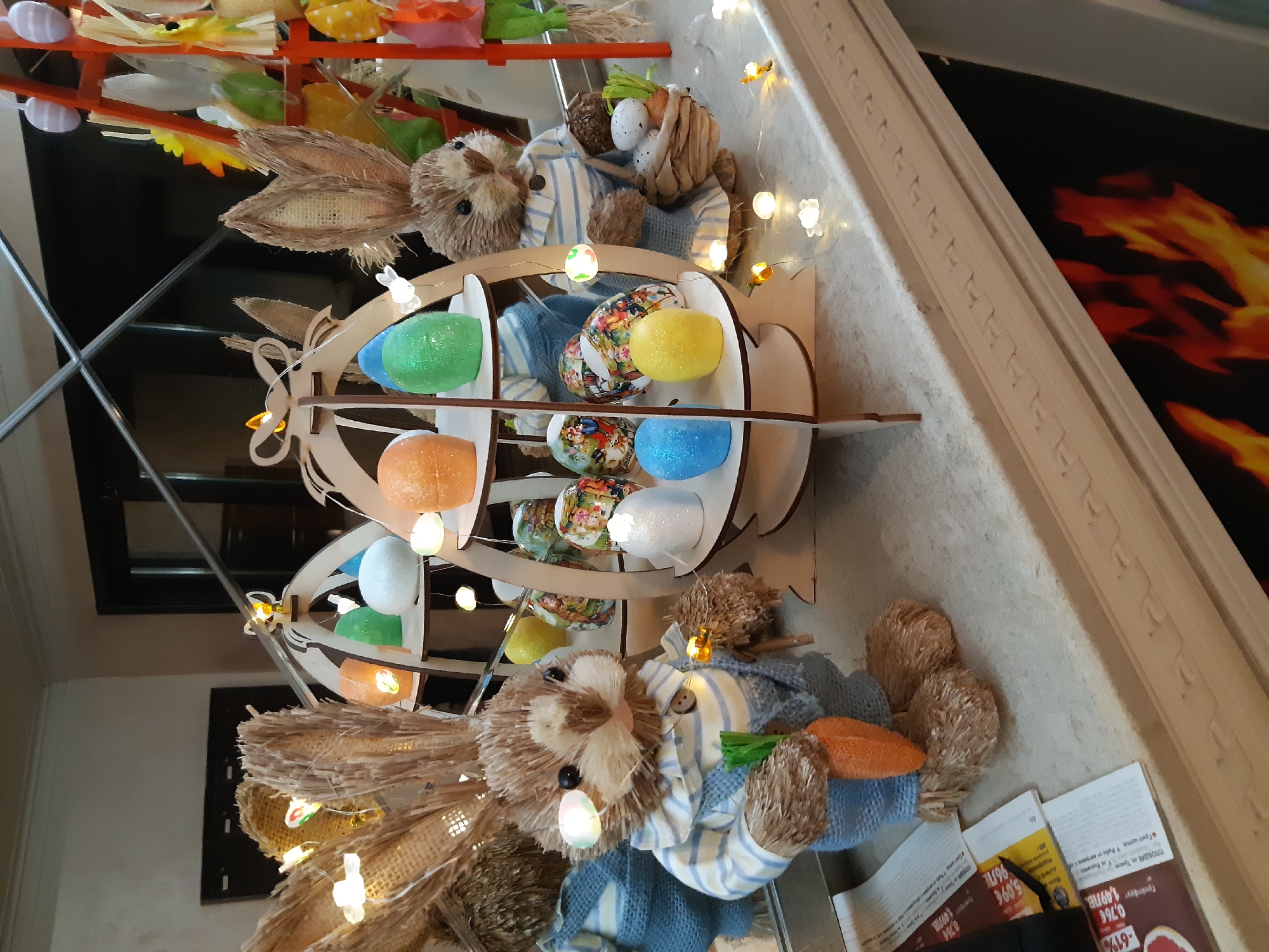 A close-up, slightly rotated image captures an Easter display. A wooden egg-shaped stand holds various colorful eggs. The stand is adorned with small lights. Adjacent to the stand are two stuffed bunny toys, one holding a carrot. The display is situated on a surface with promotional flyers visible. The background includes a mirrored surface reflecting the display, and a portion of a wall with a flame design.