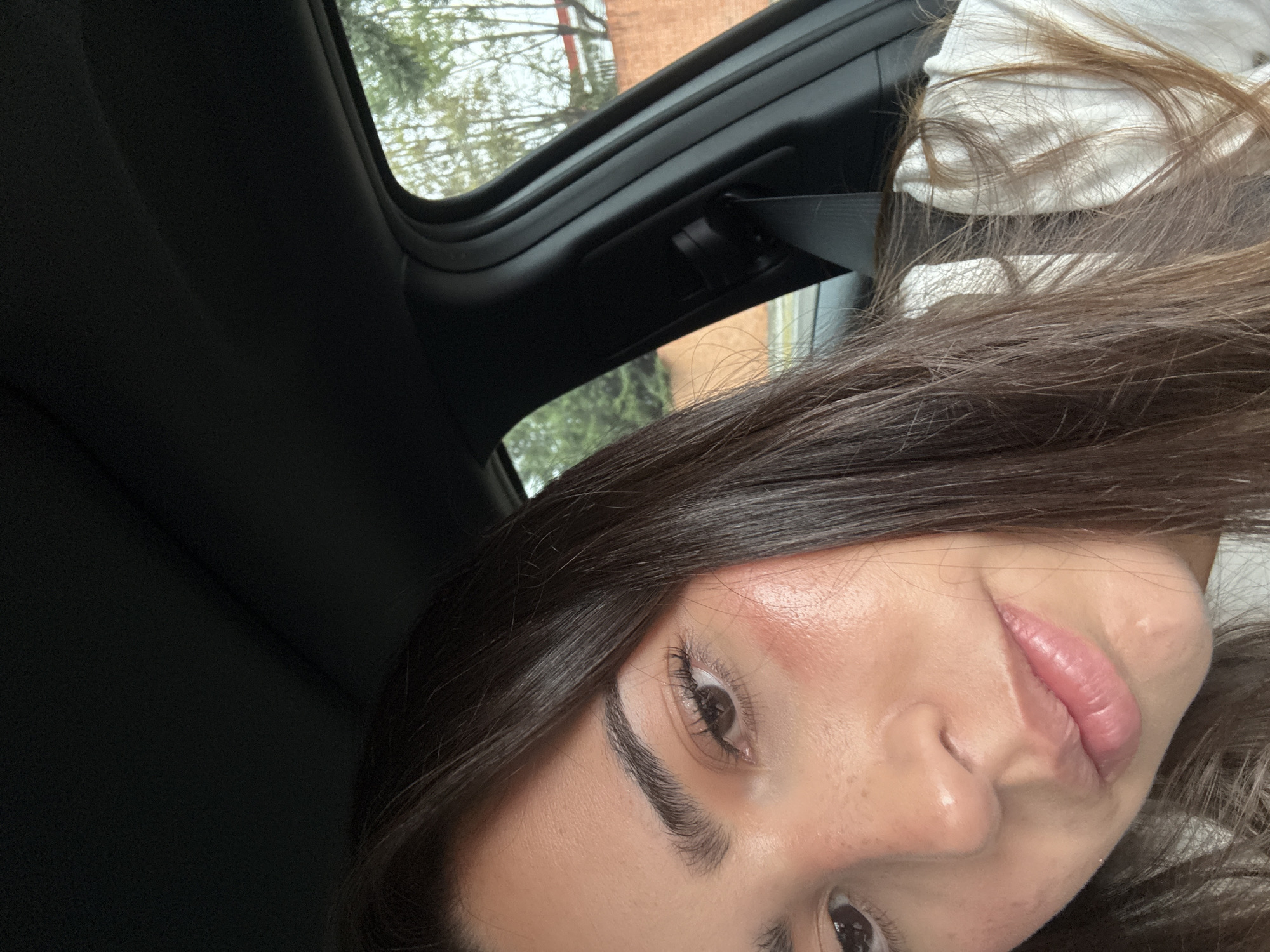 A selfie photograph of a woman with light brown hair, brown eyes, and light skin, taken from inside a vehicle. The interior of the vehicle, including the black roof and a window frame, is visible. Outside the window, there are green trees and a glimpse of a red brick building. The exact location within the city of Puebla, Mexico cannot be determined from this image.