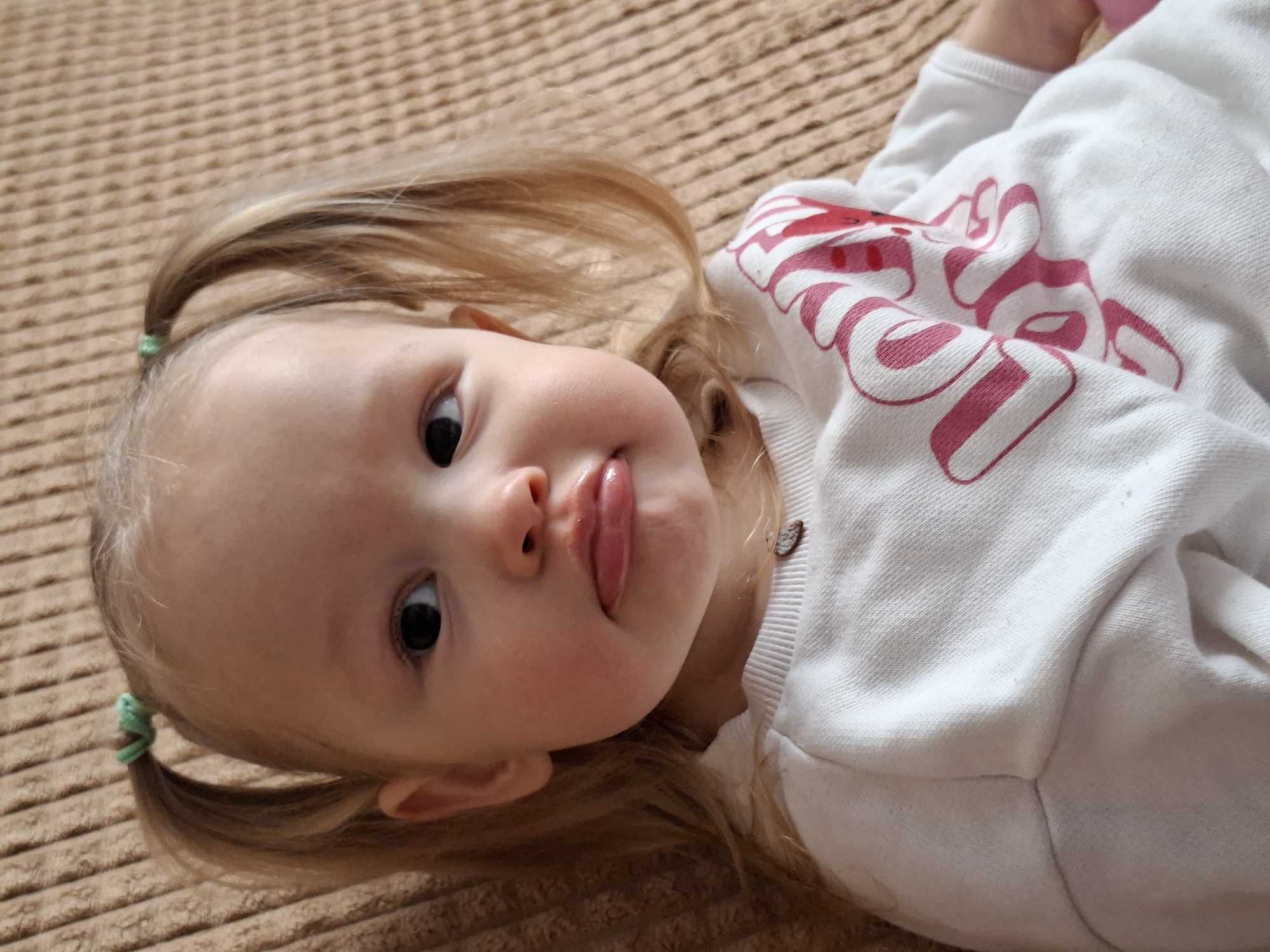 A young child, likely a toddler, is depicted in a close-up, slightly high-angle shot, lying on their back on a textured brown surface. Their head is tilted to the left, and they are looking directly at the camera with wide, dark eyes. A playful expression is conveyed by their slightly open mouth with the tongue peeking out.
The child has light-colored hair, styled into two small pigtails, each secured with a light green hair tie. Strands of hair fall around their face. They are wearing a white long-sleeved top, which features large, stylized pink text or a design across the chest. The text consists of thick, rounded block letters with outlines, partially obscured by folds in the fabric and the child's arm. Parts of the word are visible, suggesting a name or character like "SNOOPY," though it is not fully legible.
The background is a uniform, light brown textured fabric, possibly a blanket or bedspread, exhibiting a ribbed or woven pattern. The lighting appears natural, suggesting an indoor daytime setting. The image captures a candid moment, highlighting the child's innocent and curious demeanor. No specific time of day or weather conditions are discernible beyond being indoors during daylight.