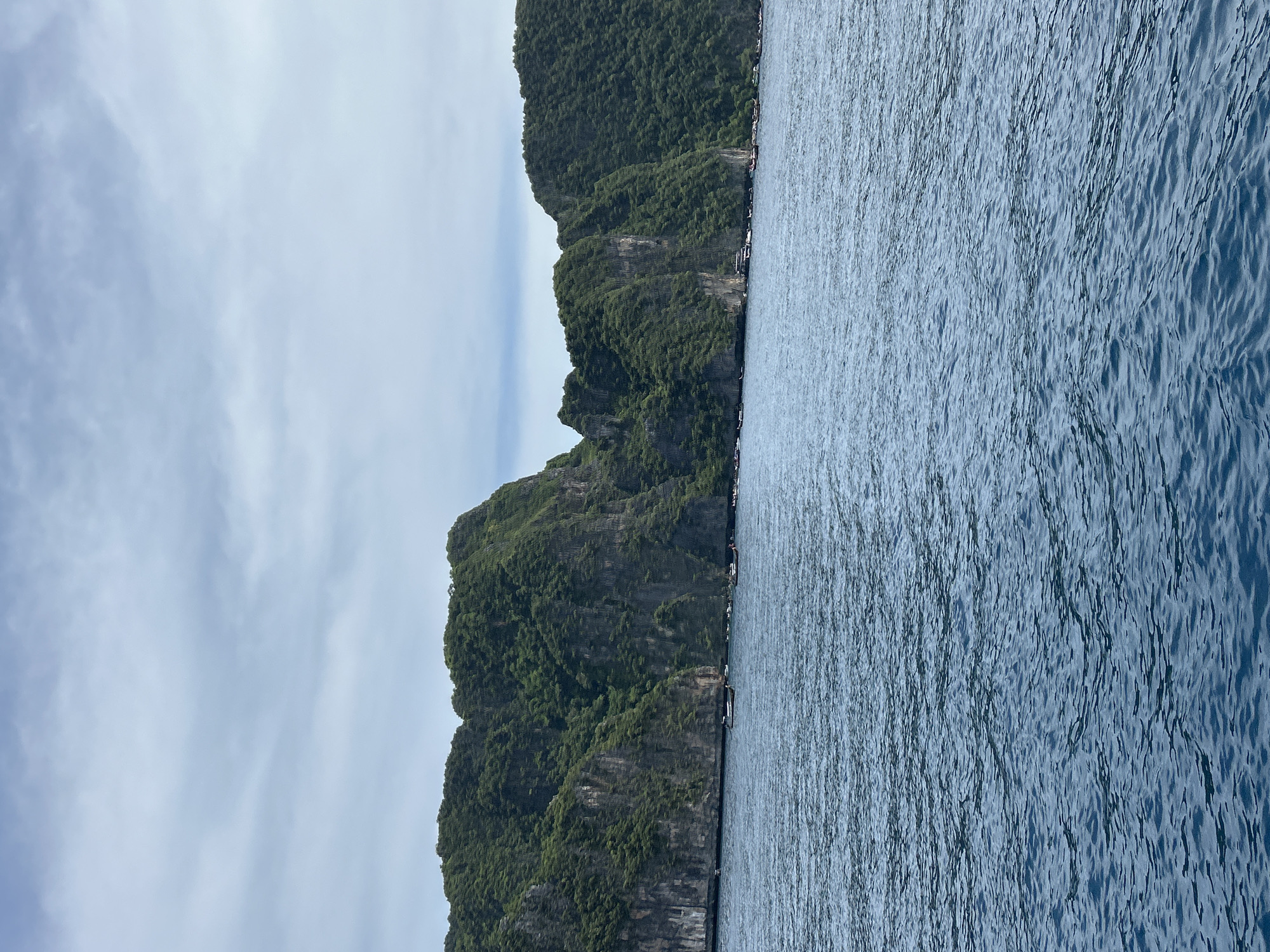 This is a medium-shot, eye-level, outdoor photograph of a coastal scene in Thailand. The foreground is dominated by choppy, dark blue water, suggesting a breezy day. In the middle ground, a long, narrow island or peninsula with steep, verdant cliffs rises from the water. The cliffs are covered in lush green vegetation, and their rugged texture is visible. Several longtail boats, typical of Thailand, are moored along the base of the cliffs. In the background, the sky is overcast with scattered light gray clouds against a pale blue expanse. The overall impression is of a natural, tropical landscape.