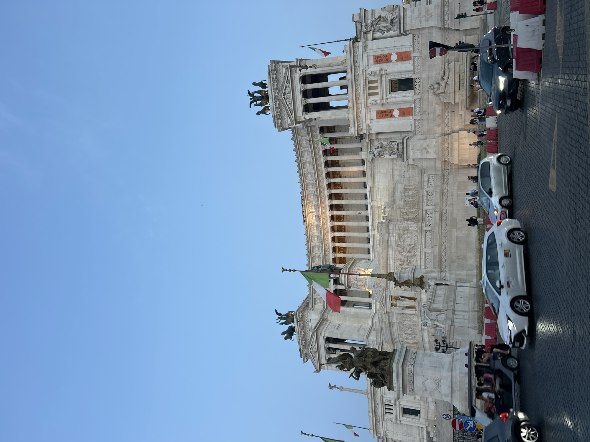 The image captures the imposing facade of the Altare della Patria, also known as the Monument to Victor Emmanuel II, in Rome, Italy. The monument, built of white marble, features grand colonnades, statues, and ornate carvings. Two Italian flags are prominently displayed on poles in front of the structure. In the foreground, a street scene unfolds with several vehicles, including a white station wagon serving as a taxi, a silver hatchback, a dark SUV, and a small electric cart carrying passengers. Traffic barriers, colored red and white, are visible on the street, indicating ongoing road work or a controlled area. Pedestrians are gathered on the sidewalks and around the monument, some appearing to be tourists. The sky above is a clear, pale blue, suggesting it is either early morning or late afternoon. The architectural style of the monument and the presence of Italian flags strongly suggest the location is Rome.