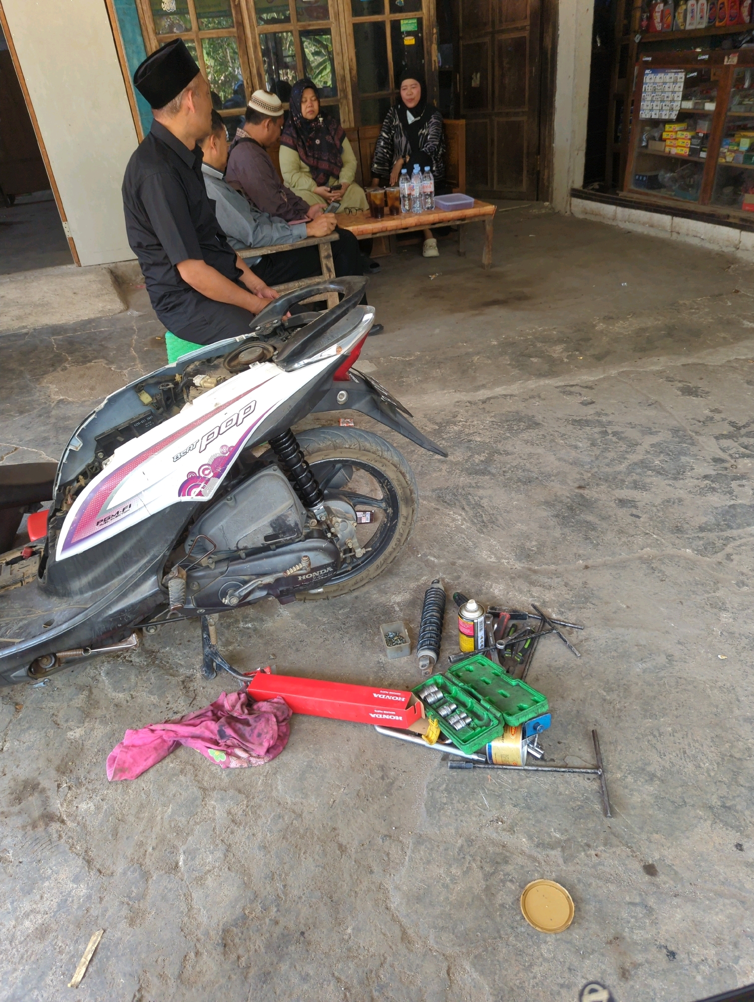 In Kedungdowo, Indonesia, a man wearing a black shirt and peci sits on a green stool next to a partially dismantled motorcycle. Several people are gathered around a wooden table behind him, with one woman in a headscarf holding a mobile phone. Tools and spare parts for the motorcycle are scattered on the concrete ground in the foreground. The scene appears to be outside a building with a storefront that has shelves stocked with various items.