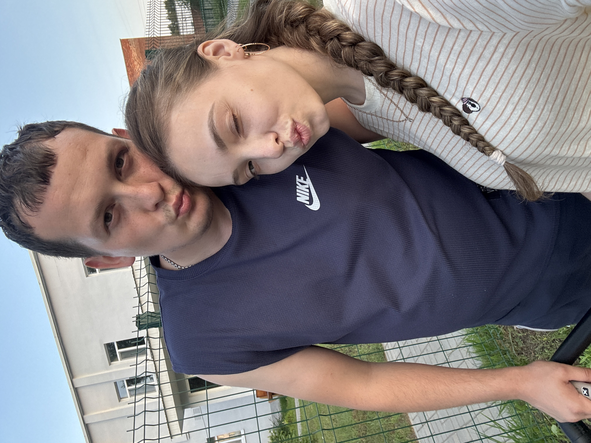 A young Caucasian couple is captured in a selfie taken outdoors, with a building and a fence visible in the background. The man, on the left, is wearing a dark blue sleeveless Nike shirt. He has short brown hair and is puckering his lips towards the camera. The woman, on the right, has long brown hair braided down her back. She is wearing a light-colored, horizontally striped shirt with a small embroidered patch on the chest. Her right eye is closed as she also puckers her lips. The setting appears to be a warm day, with a clear sky. The man's left hand is visible holding onto a dark object, possibly a bicycle handlebar, with a white grip. The location of Noviy, Russia is mentioned in the prompt, but there are no specific visual cues in the image to confirm this exact location. The overall atmosphere of the image is casual and affectionate.