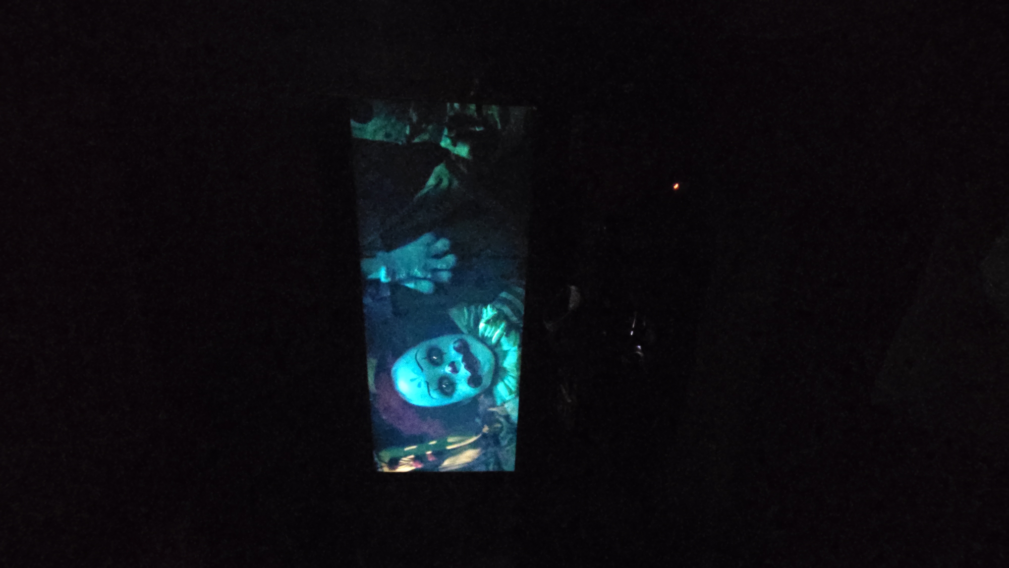In a dark room, a television screen displays an image of Pennywise the Clown from the movie "It." The clown has a pale face with blueish-green undertones, red lips, and white makeup around its eyes and mouth. It wears a ruffled, multi-colored costume. To the right of the clown, a hand in a white glove is visible, reaching out. The background of the image on the screen is dark and appears to be a dimly lit, possibly outdoor, environment with some green foliage. The room itself is mostly black, with only the glow of the television screen providing illumination. A small red light, possibly from a device, is visible in the lower right portion of the image, indicating the general location of Il'inskiy, Russia.