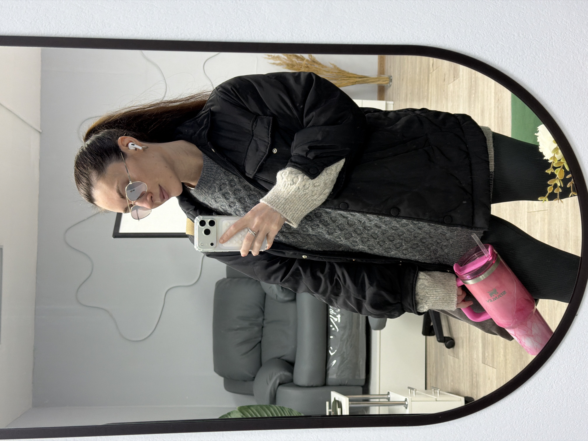 A woman is reflected in a mirror, presumably taking a selfie. She has brown hair pulled back in a ponytail, and is wearing gold-rimmed glasses and an AirPod in her left ear. She is wearing a black puffer jacket over a grey patterned sweater and dark leggings. She is holding a white smartphone in her left hand. Her right hand holds a pink Stanley tumbler with a straw. A gray chair and a set of white drawers with silver rails are visible in the background. A picture frame hangs on the wall. The location could be Perm, Russia based on text partially visible on the chair.