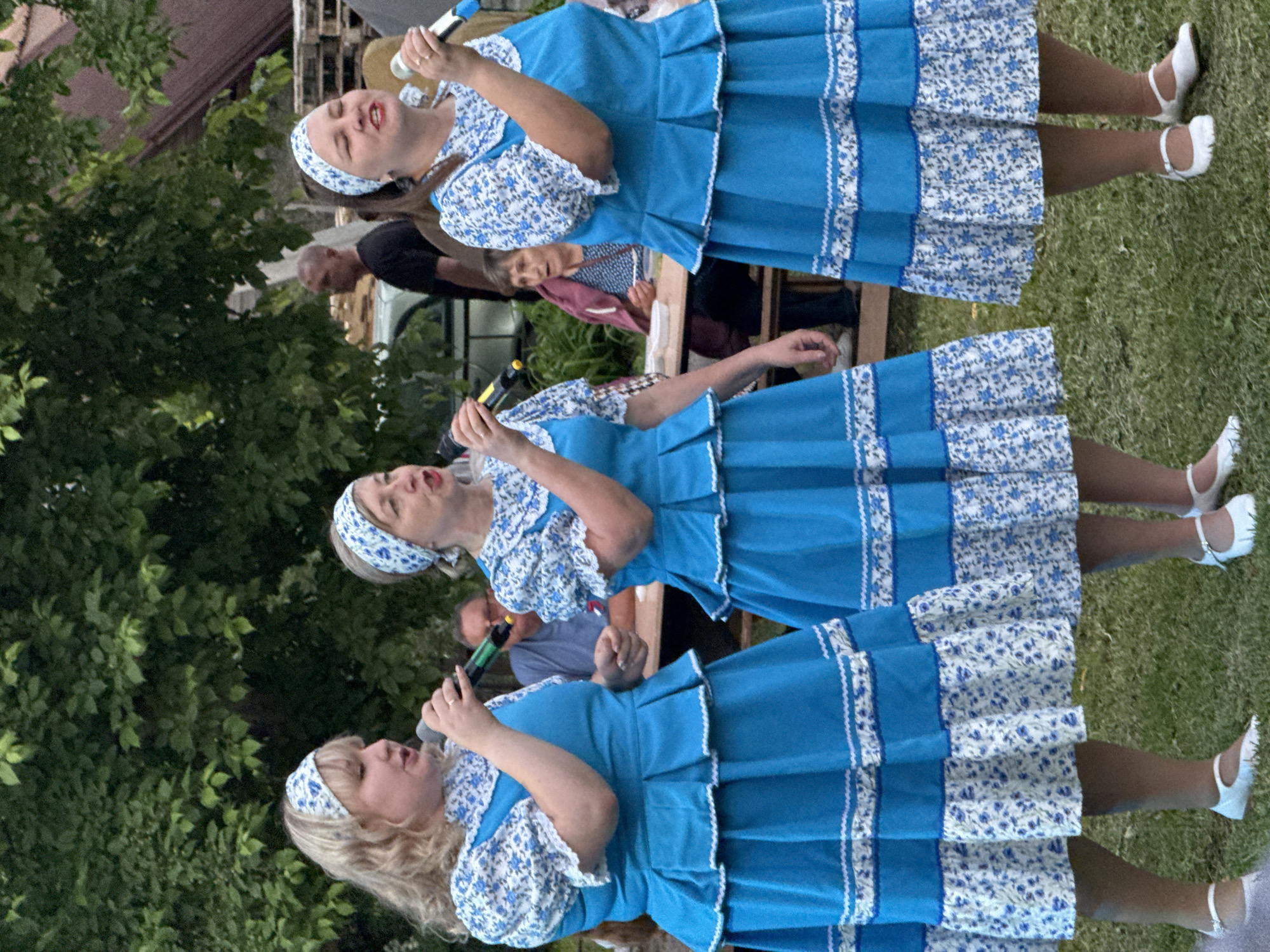 Three women, dressed in matching blue and white folk costumes with floral patterns and headbands, are singing into microphones. They stand on a grassy area, in front of trees and some outdoor structures. The woman on the left has blonde curly hair, the middle woman has graying blonde hair, and the woman on the right has dark hair. In the background, other people are visible, including a man in a black t-shirt and a woman seated at a table. The scene appears to be an outdoor performance or gathering. This event is taking place in the village of Kukui, Russia.