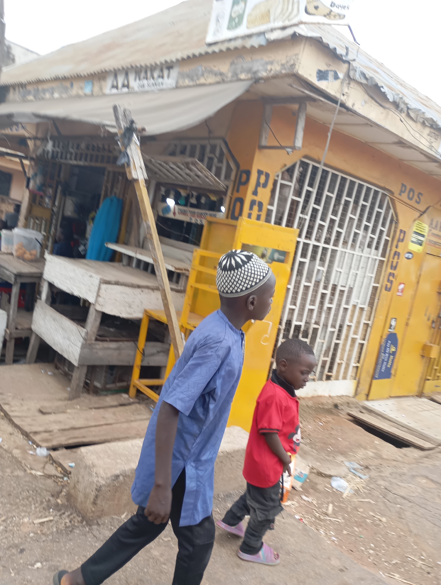 The image depicts a street scene in Shendam, Nigeria. Two young boys are walking on a dirt road in front of a yellow building. The boy on the left, wearing a blue tunic and black pants, has a white and black patterned kufi on his head. The boy on the right, wearing a red t-shirt with a Mickey Mouse graphic and gray pants, has on pink sandals. The yellow building appears to be a shop or business, with barred windows and signage that is partially obscured. Part of a weathered awning is visible above the entrance. Wooden stalls and debris are scattered on the ground around the building. The overall environment suggests a bustling market or commercial area.