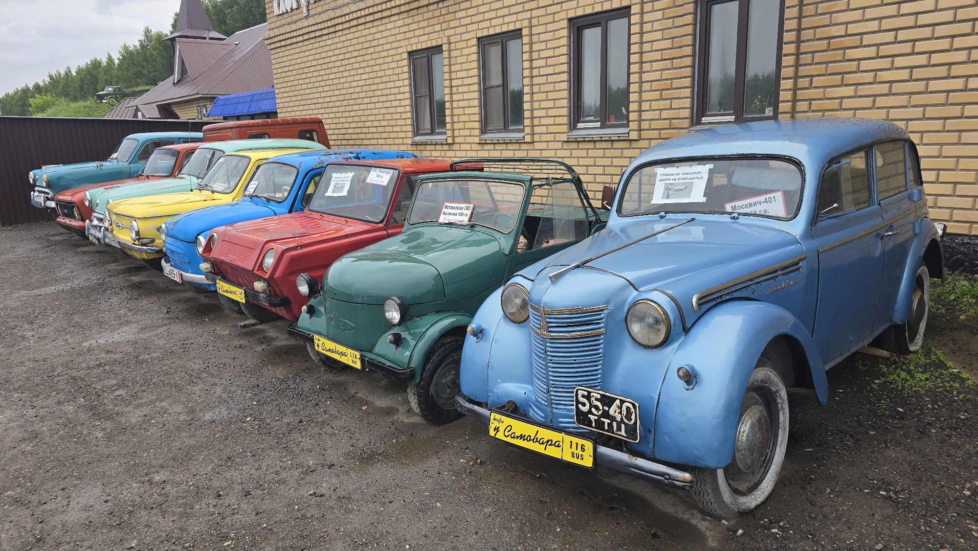 In Bol'shoi Mashlyak, Russia, a line of vintage cars is parked on a gravel surface in front of a brick building. The cars are brightly colored, including a teal sedan, an orange sedan, a yellow sedan, a blue sedan, a red microcar, and a green microcar. The building has a brick facade with several windows. In the background, there is a wooden fence and trees. One of the cars, a blue sedan, has a license plate that reads "55-40 TTU". Another car, a green microcar, has a sign that says "Москвич-401" (Moskvich-401).