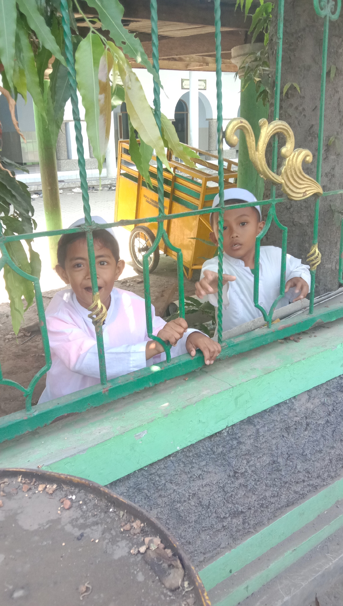 Two young boys, dressed in white traditional Islamic clothing and skullcaps, are looking through a green metal fence. The boy on the left, with a light pink outer garment, has a wide, open-mouthed expression, appearing surprised or excited. He is leaning on the fence with both hands. The boy on the right, wearing a plain white outer garment, is reaching out a hand towards the fence, his expression more neutral. Behind them, partially visible through the fence, is a yellow cart with green trim, possibly a market cart or a utility vehicle. The background suggests an outdoor setting with some vegetation and a building with white arches and a mosque-like structure. The scene appears to be in an urban or semi-urban area, possibly a public space like a schoolyard or a street in Karang Anyar, Indonesia. The lighting suggests a daytime scene, likely under direct sunlight.