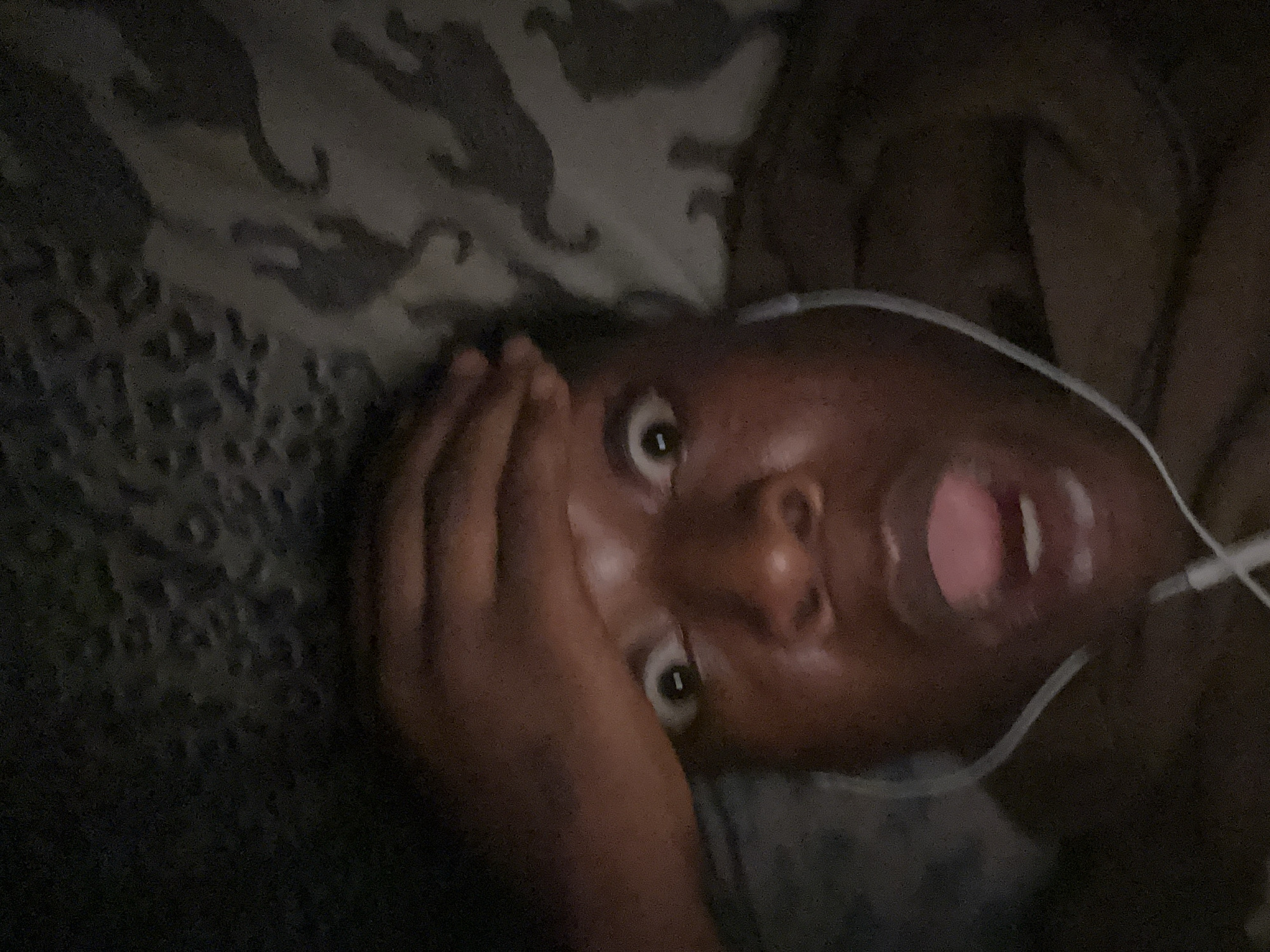 A person with dark skin is lying down with their head turned upward, mouth open in a gasp or yell, and eyes wide. Their hand is partially covering their face. They are wearing white earphones with the cord visible. The background appears to be a patterned bedspread or blanket. The location and city are unknown.