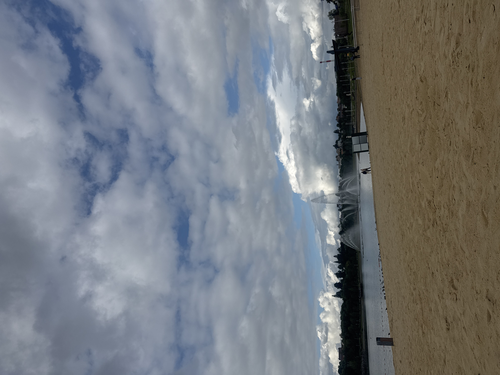 The image depicts a beach area bordering a body of water with a large fountain in the distance. The foreground is dominated by a sandy beach. In the middle ground, the water stretches across the frame, with the fountain spraying water high into the air. The background shows a tree line with buildings and structures visible beyond. The sky is filled with dramatic clouds. A few people can be seen walking on the beach and near the water's edge. The location is identified as the embankment of the "Beloe Ozero" (White Lake) in Ufa, Russia.